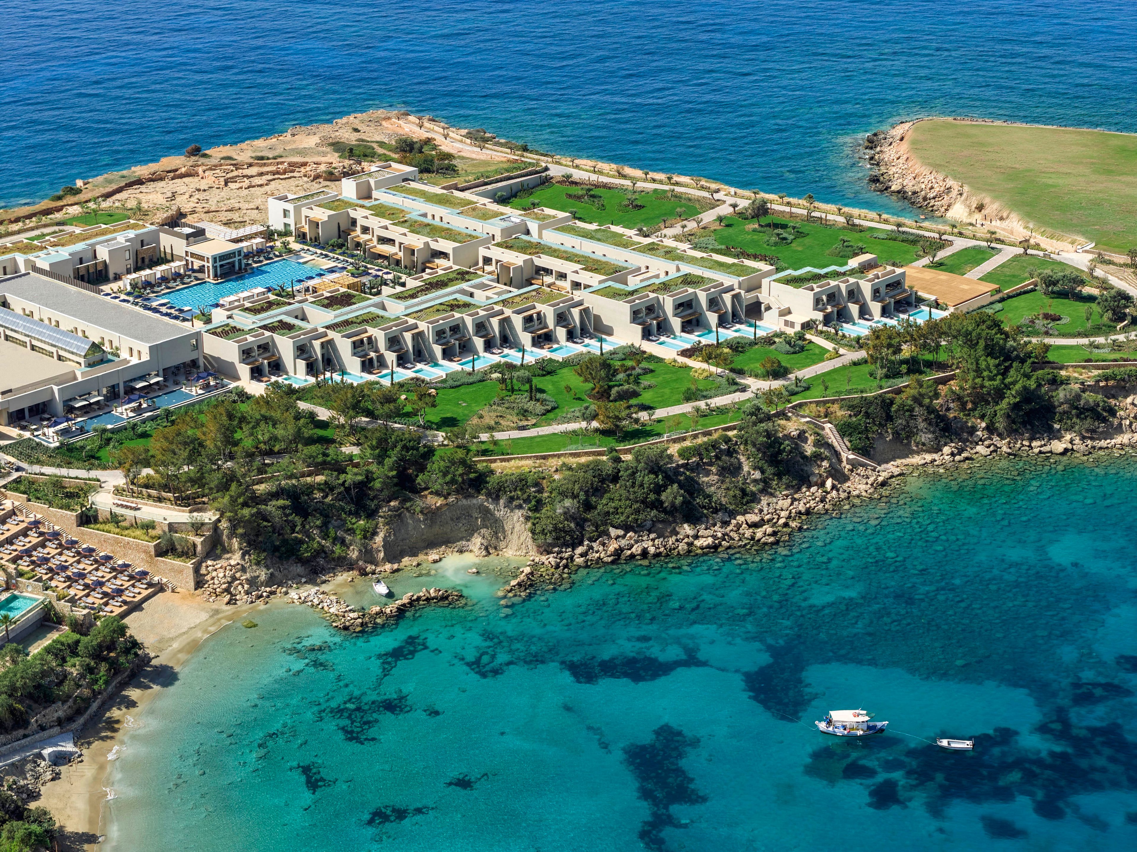 A modern luxury resort stretches across a rocky peninsula surrounded by clear turquoise sea. The complex features multiple pools, terraced suites, and manicured green lawns overlooking the coastline. Sun loungers line the sandy beach area while small boats float in the calm bay. The aerial perspective highlights the contrast between the bright buildings, lush landscaping, and deep blue ocean.