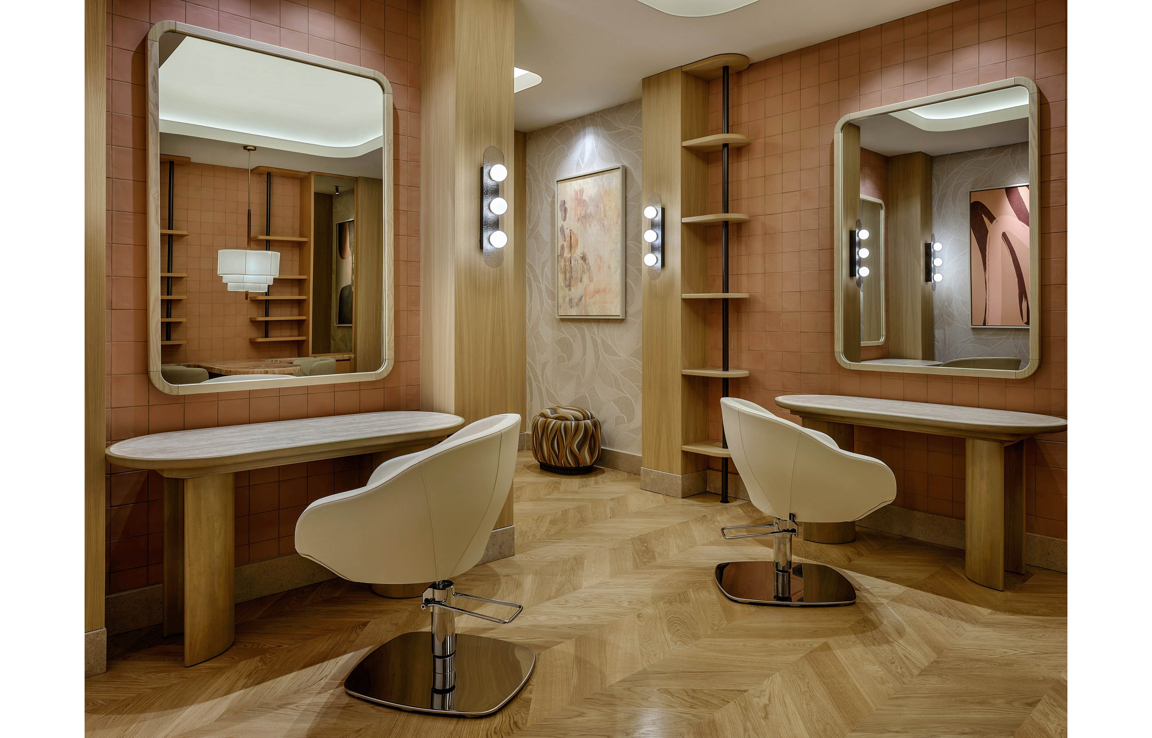 Contemporary salon interior featuring two white styling chairs facing large rectangular mirrors. The space is designed with warm wood tones, herringbone flooring, and soft pink tiled walls. Built-in shelves, wall sconces, and decorative artwork add a refined, minimalist touch. The overall atmosphere feels calm, luxurious, and inviting for beauty or hair services.