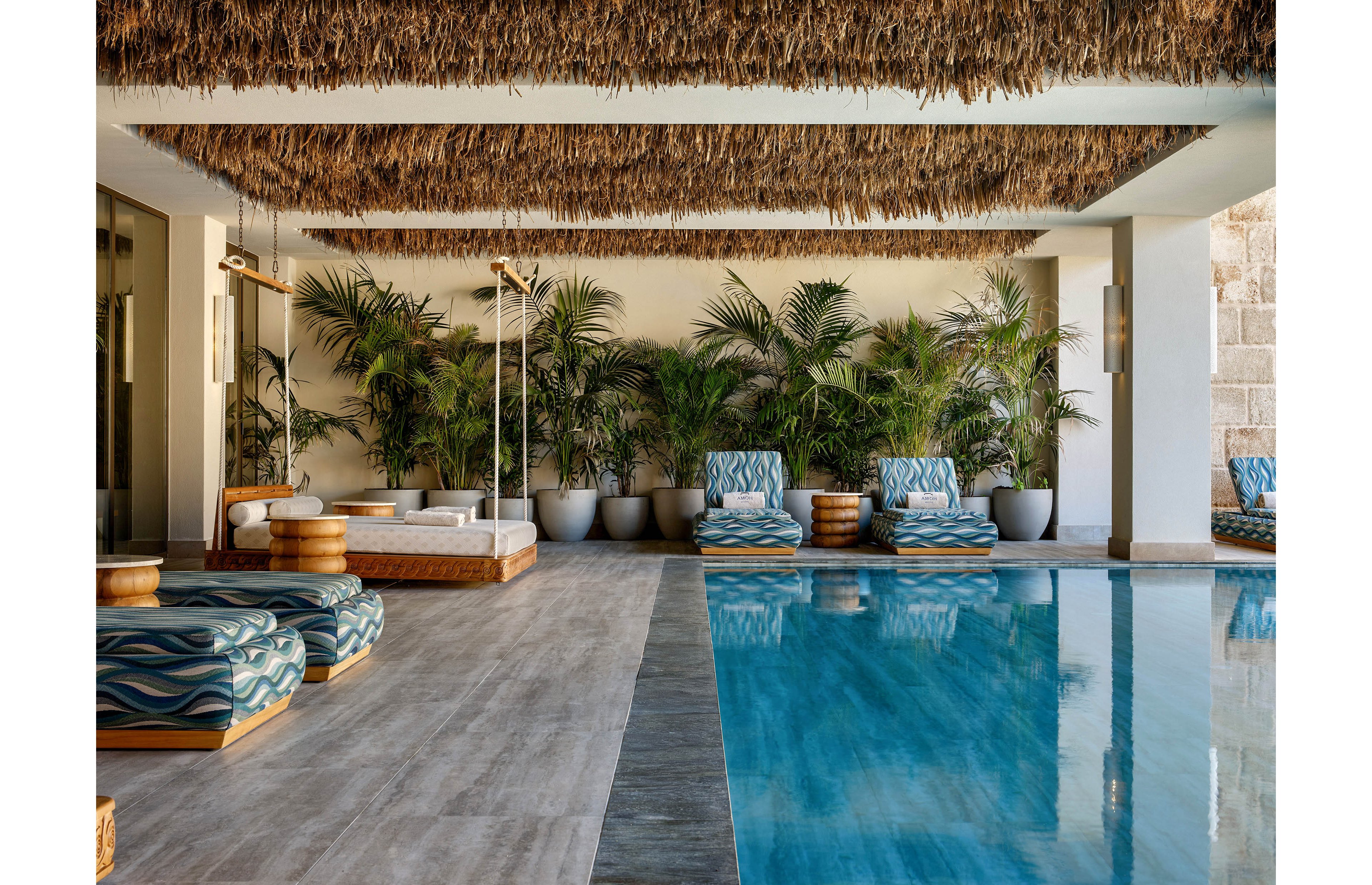 A stylish indoor pool area features turquoise water beside a row of cushioned lounge chairs. The seating has patterned blue upholstery and wooden bases, arranged along a wall filled with potted tropical plants. A thatched ceiling and neutral stone flooring create a warm, natural atmosphere. The space appears calm and inviting, ideal for relaxation in a resort or spa setting.