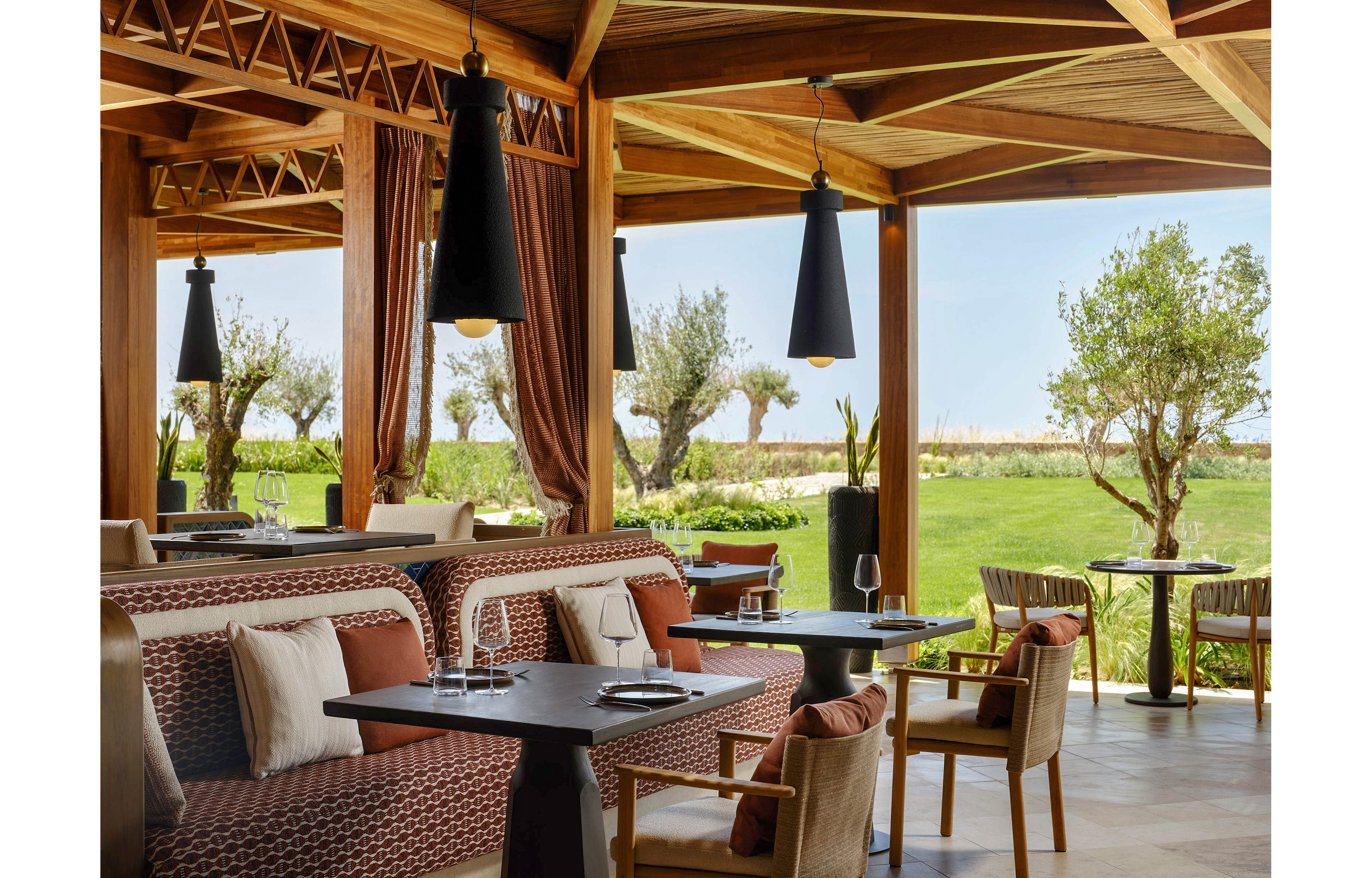 Upscale outdoor restaurant seating area with modern wooden architecture and woven upholstered booths. Black pendant lights hang above dark tabletops set with plates and empty wine glasses. Large open sides reveal a lush green lawn with trees and a bright sky in the background. The scene suggests a relaxed, elegant dining experience in a resort or garden setting.
