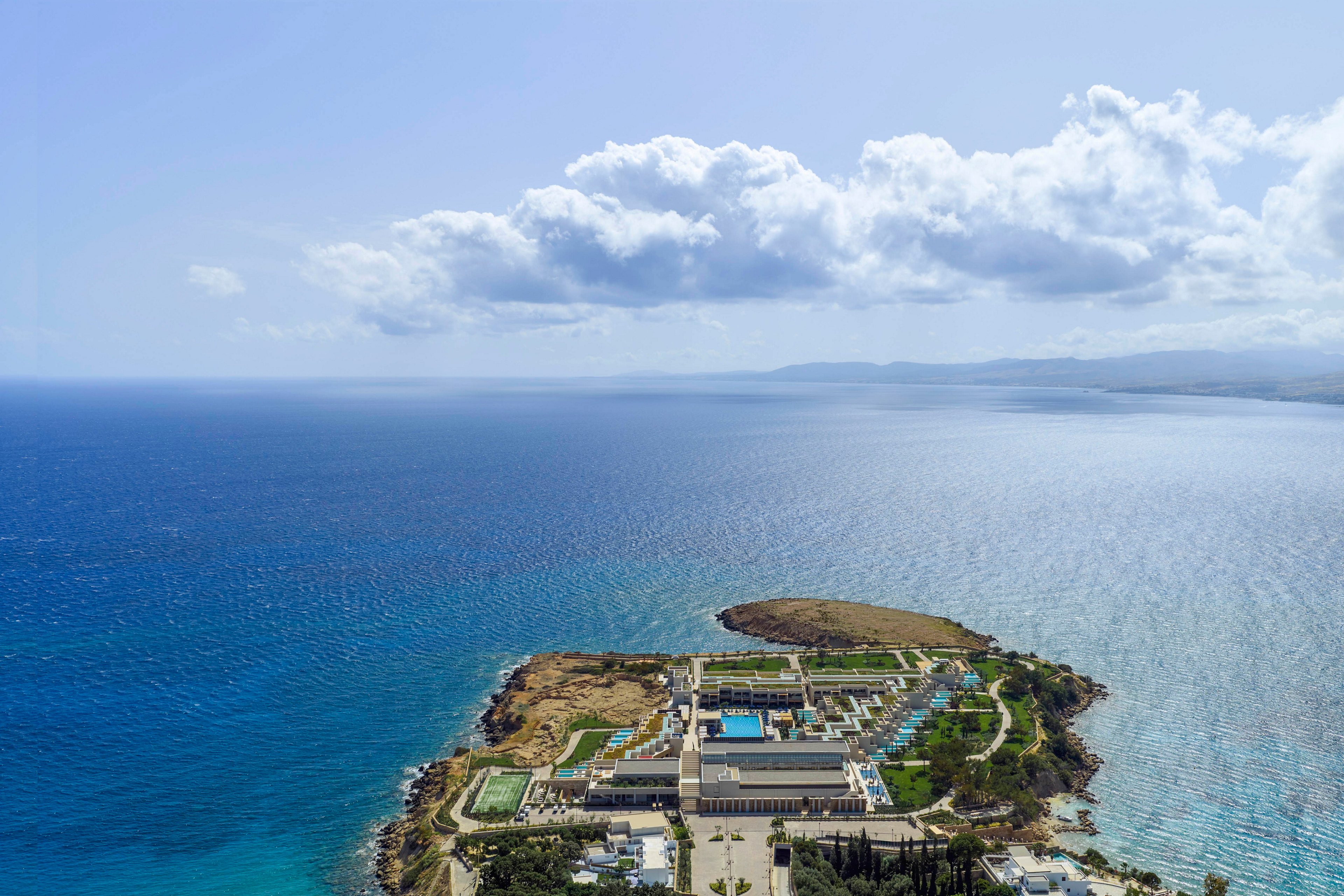A large resort complex sits on a narrow peninsula surrounded by expansive blue ocean. The aerial perspective shows multiple buildings, pools, and landscaped areas stretching toward the water. Calm seas and a distant coastline under a partly cloudy sky create a serene vacation atmosphere. The image emphasizes the resort’s isolated location and panoramic seaside views.