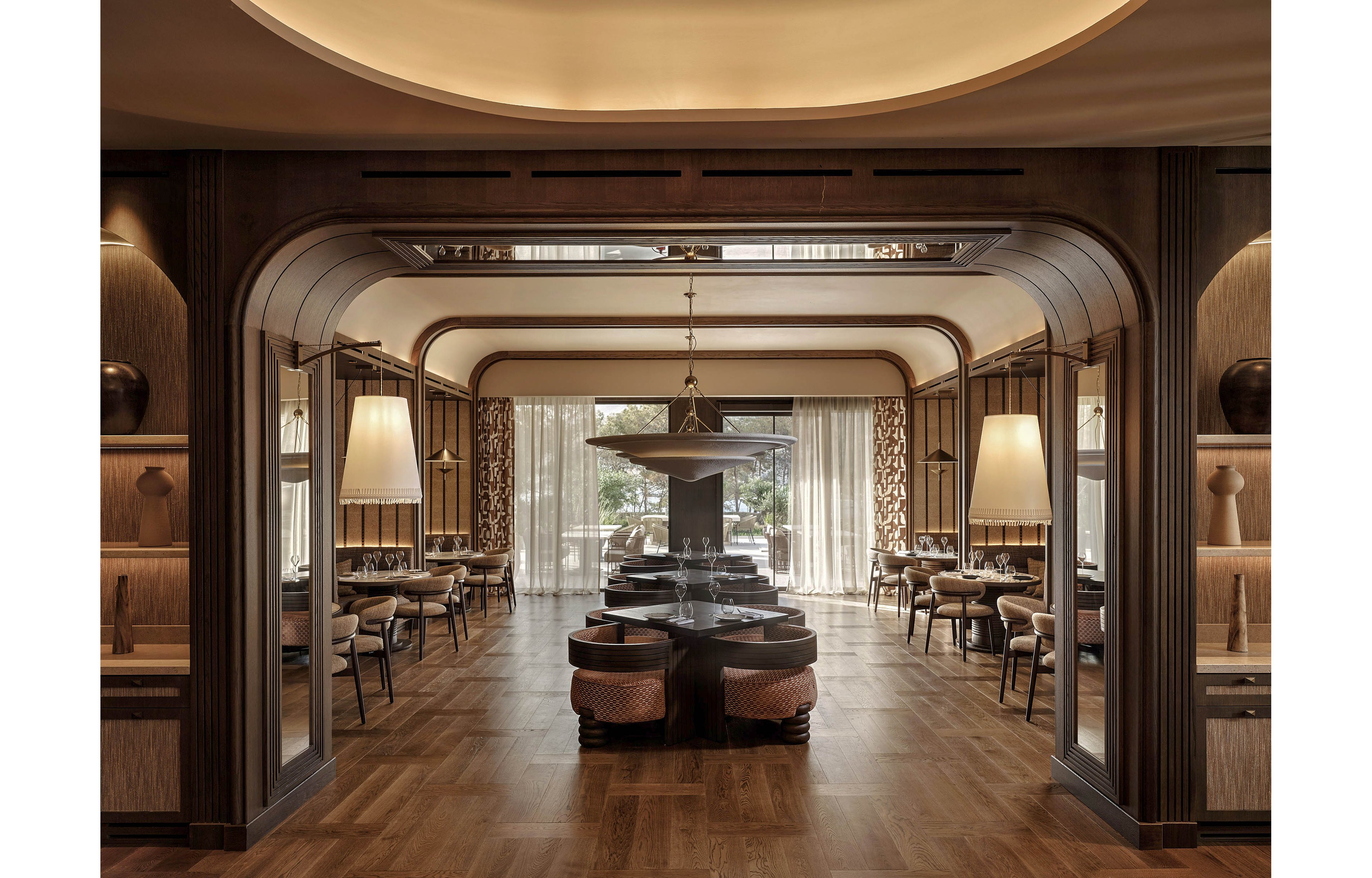 Upscale restaurant interior featuring a central seating island framed by dark wood arches. Soft pendant lighting and large windows with sheer curtains create a warm, inviting atmosphere. Dining tables and chairs line both sides of the room, arranged symmetrically for a refined look. Neutral tones, patterned drapes, and polished wood flooring emphasize a modern yet classic design aesthetic.