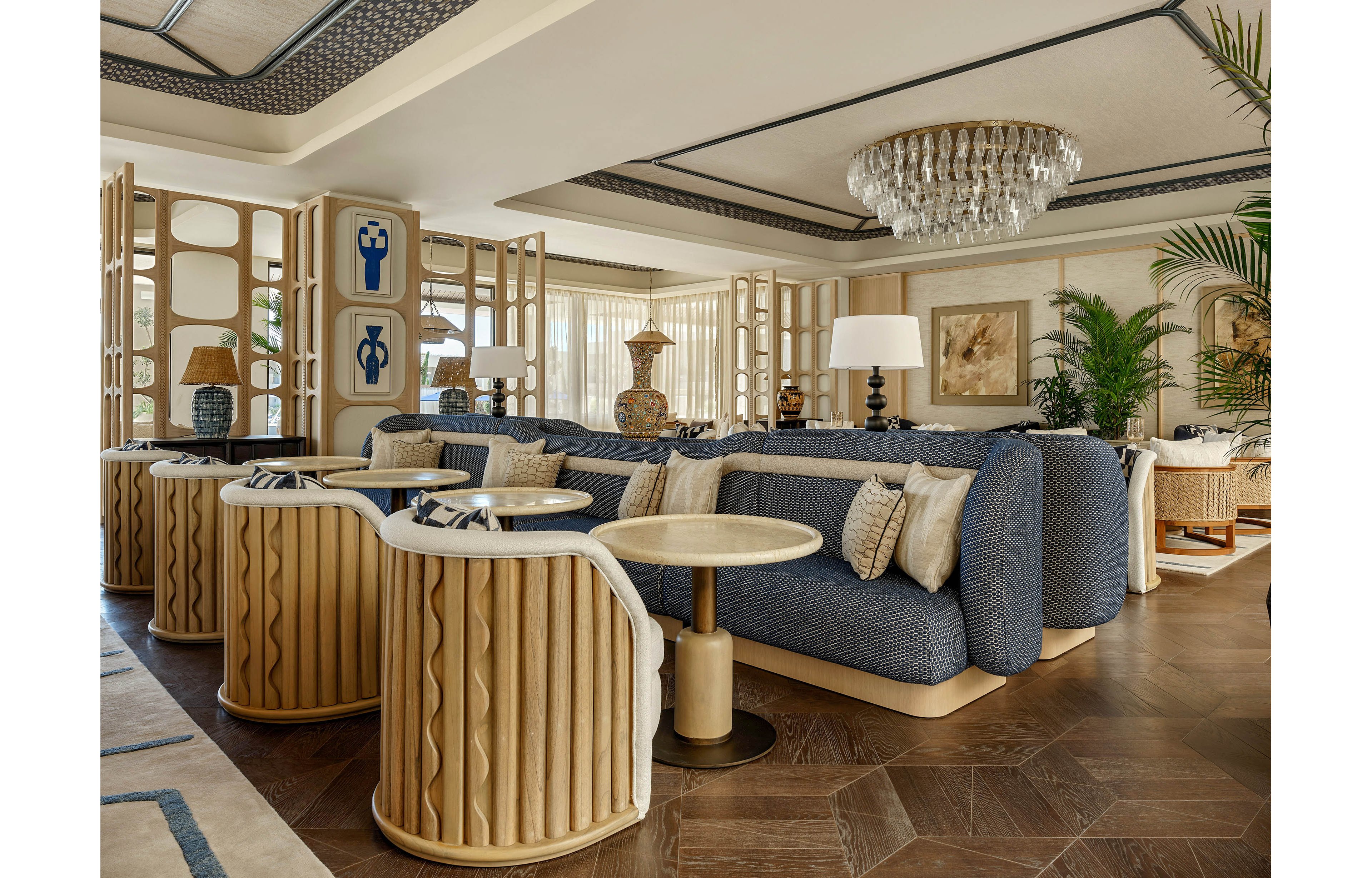Upscale lounge area featuring curved blue sofas and beige accent chairs arranged around small round tables. The space is decorated with sculptural vases, patterned cushions, and decorative wooden room dividers. Large windows with sheer curtains allow natural light to soften the warm neutral color palette. A crystal chandelier and modern table lamps add an elegant, sophisticated ambiance to the interior.