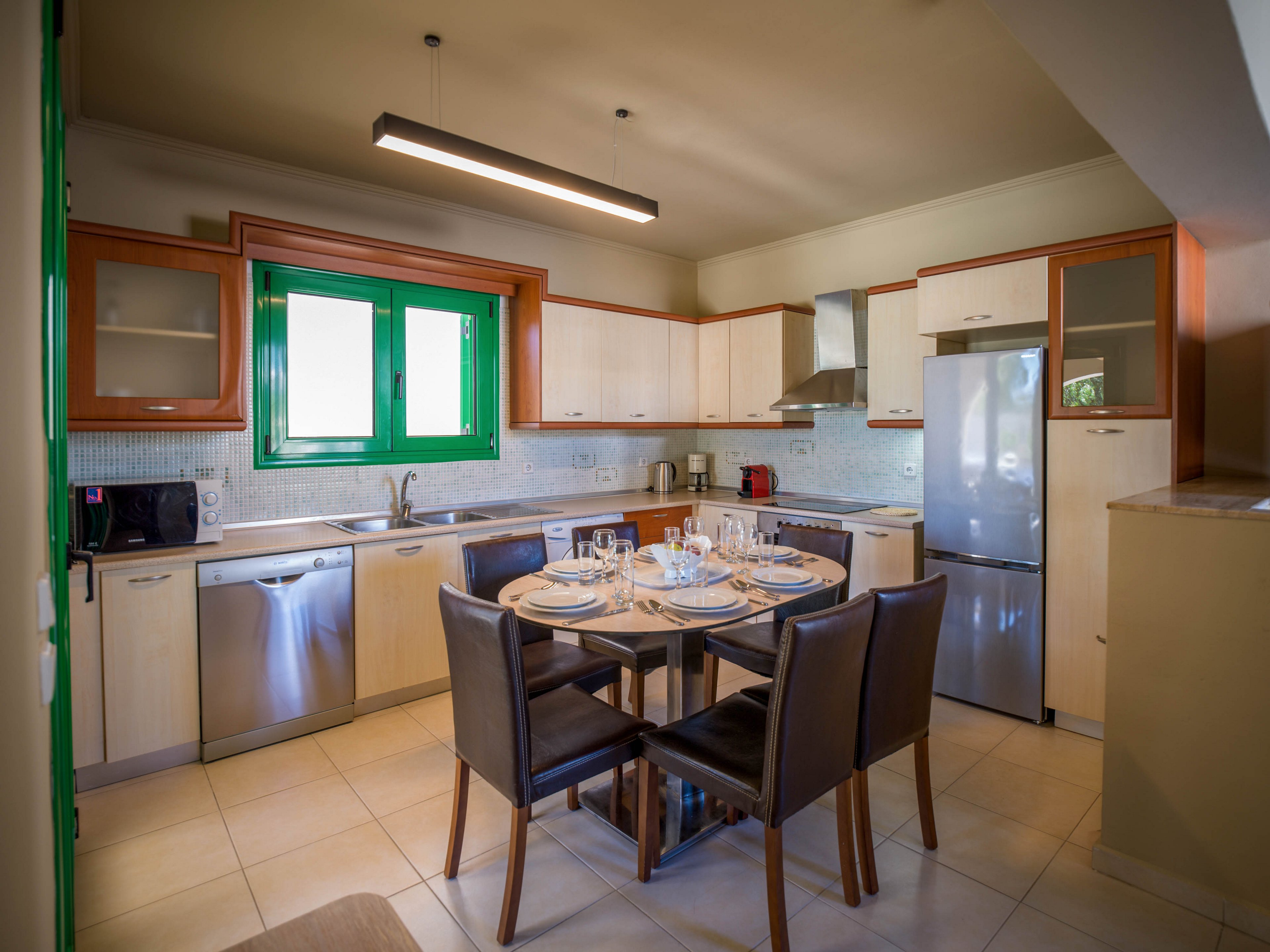 A contemporary kitchen features light cabinetry, stainless steel appliances, and a central round dining table set for four. The space includes a refrigerator, dishwasher, oven, microwave, and small countertop appliances arranged neatly along the walls. A bright green-framed window adds a pop of color and natural light to the neutral-toned room. The overall scene suggests a clean, ready-to-use kitchen in a modern home or vacation rental.