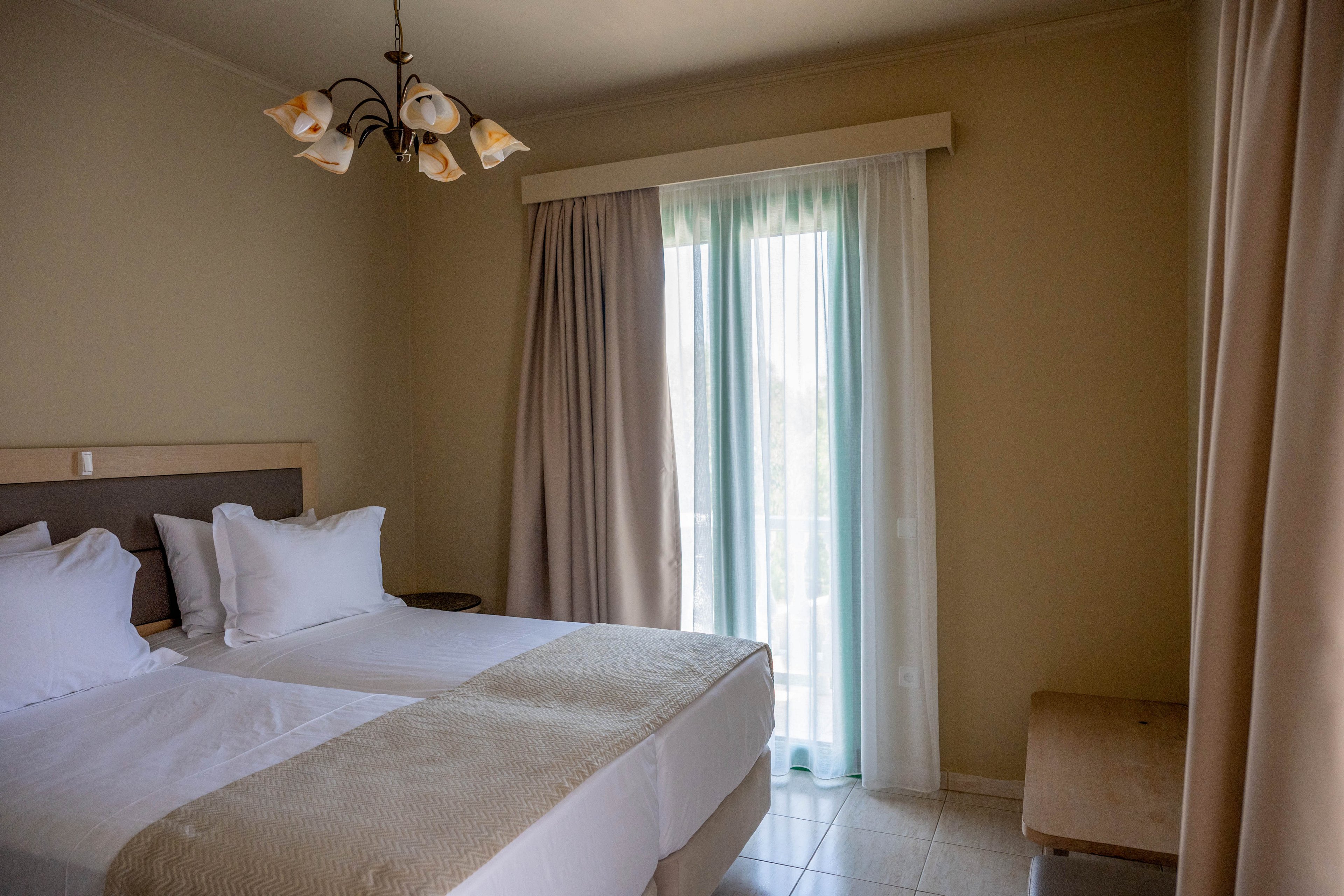 A tidy hotel room features two neatly made twin beds with white linens and beige throws. Soft natural light filters through sheer curtains and heavier drapes covering a tall window or balcony door. A simple wooden bench sits against the wall, and a ceiling light fixture hangs above. The neutral color palette and minimal decor create a calm, inviting atmosphere.