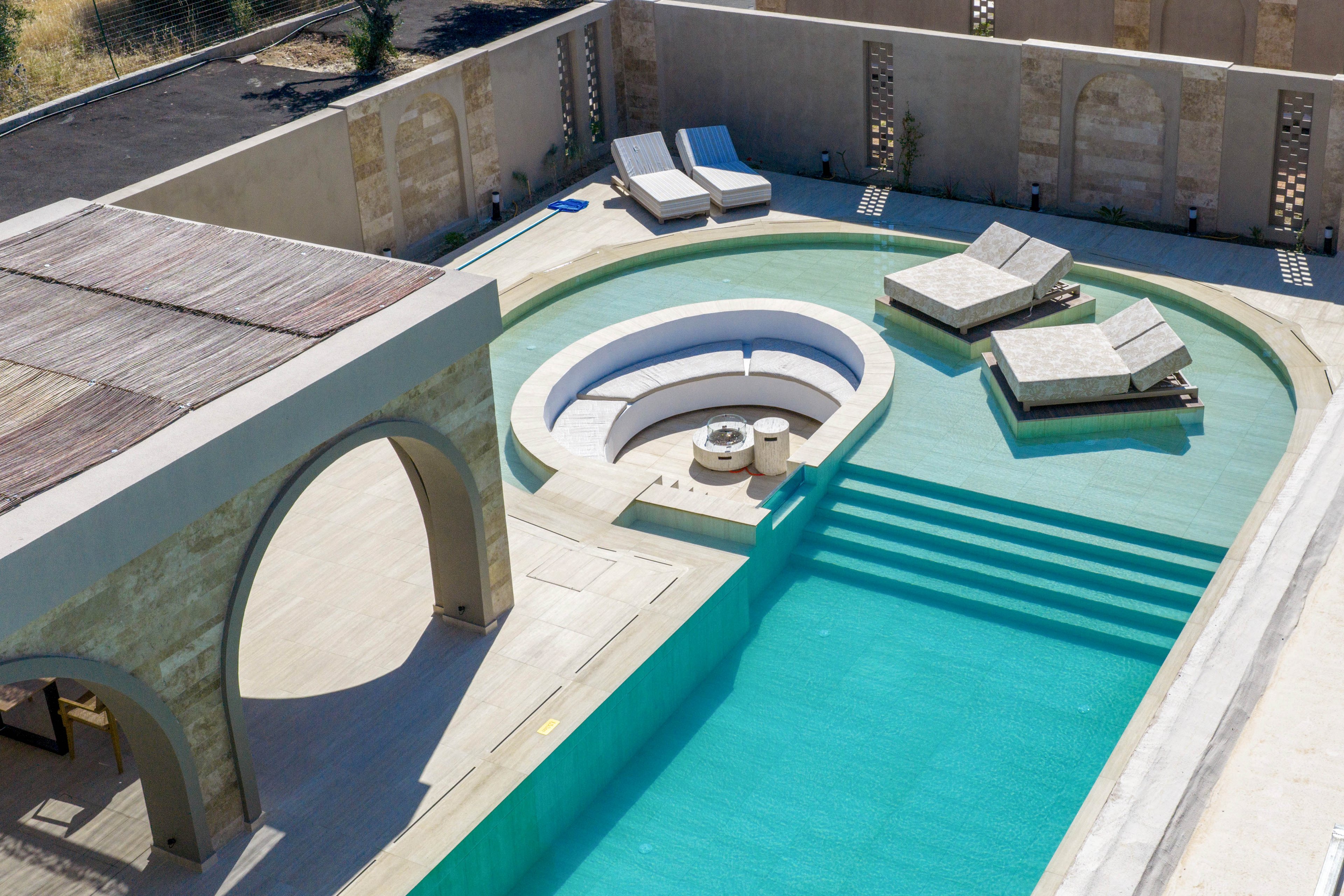 A modern luxury swimming pool features an elegant circular sunken seating area at its center. Several cushioned sun loungers are arranged on platforms partially over the water, inviting relaxation. The surrounding architecture has a minimalist, Mediterranean-inspired design with light stone walls and arched structures. Bright turquoise water and neutral beige tones create a calm, upscale resort atmosphere.