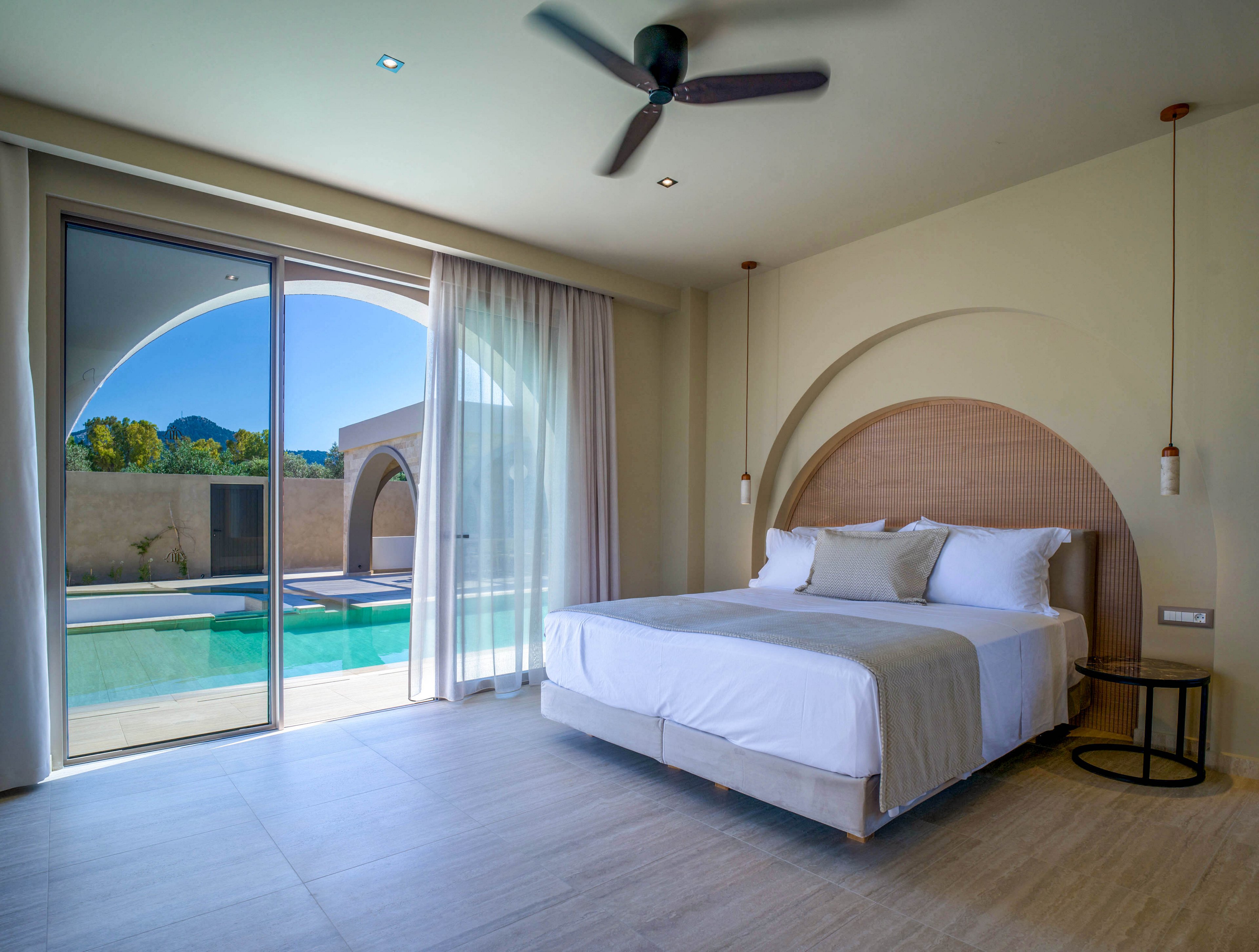 Contemporary bedroom with a neatly made double bed faces large sliding glass doors. Outside, a private swimming pool and arched architectural details are visible under a clear blue sky. The interior features neutral tones, a circular accent wall, and minimalist pendant lighting. Sheer curtains, a ceiling fan, and clean lines create a calm, airy atmosphere.