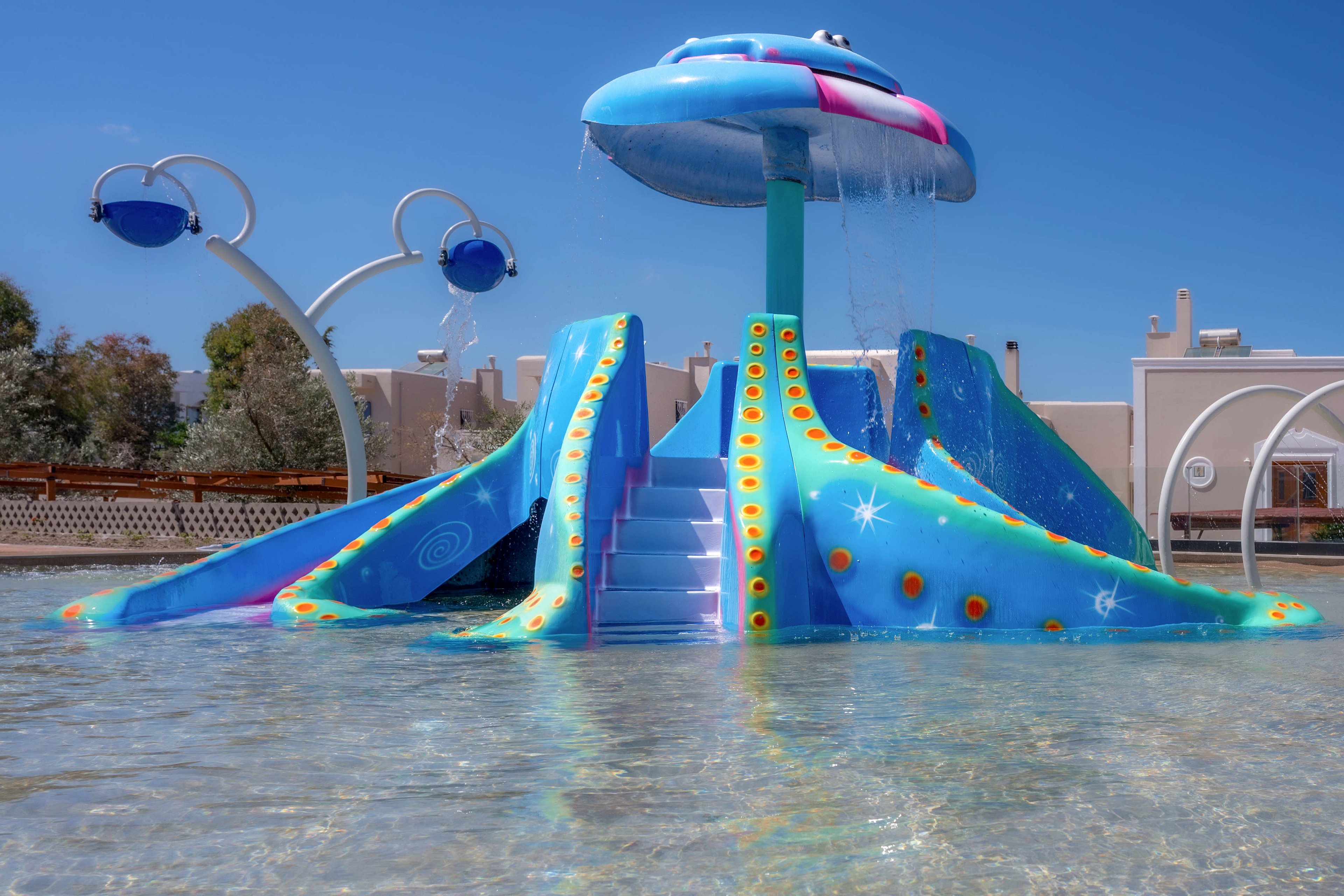 A bright, UFO-shaped waterslide structure stands in a shallow splash pool under a clear blue sky. The slide features multiple ramps, a central staircase, and playful star and bubble patterns in blue, orange, and pink. Water cascades from the top of the dome, creating a gentle fountain effect. Modern buildings and additional water play features are visible in the background, suggesting a family-friendly water park setting.
