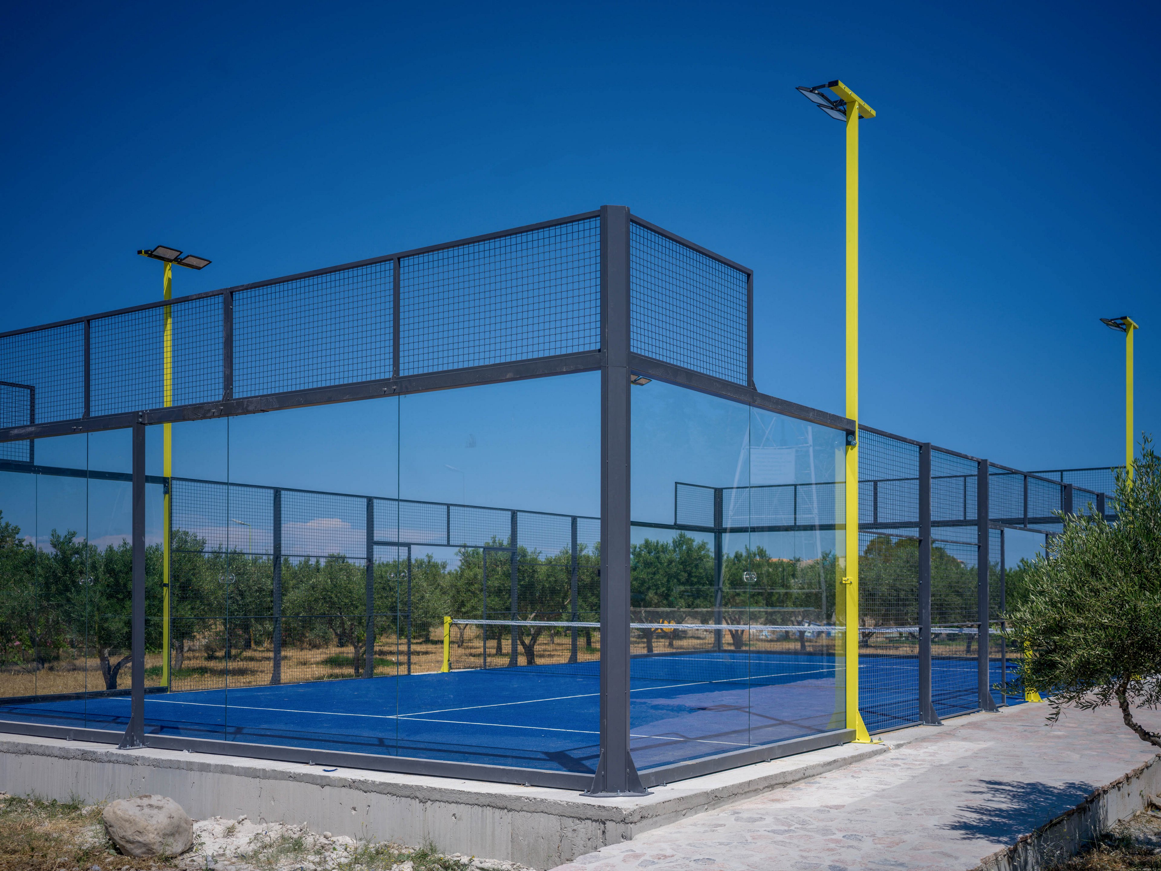 An outdoor padel court with a bright blue playing surface is enclosed by glass and metal fencing. Tall yellow light poles rise above the structure under a clear blue sky. The court is situated in a rural setting with trees and dry grass surrounding the area. A paved walkway leads up to the court, emphasizing its recreational purpose in a natural landscape.