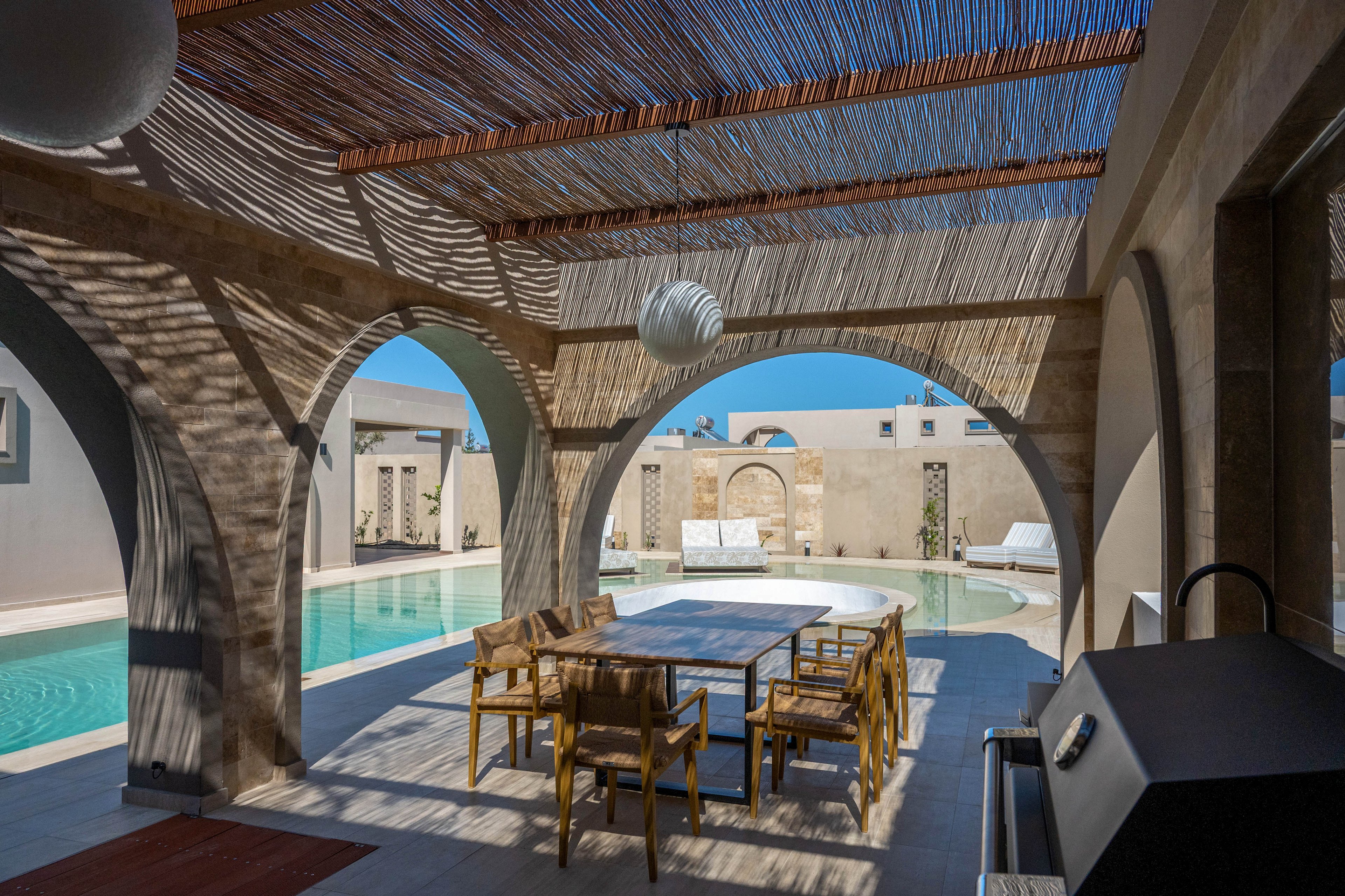 An outdoor patio with arched openings overlooks a clear blue swimming pool. A wooden dining table with several chairs sits under a slatted pergola that casts patterned shadows on the floor. Lounge chairs and sunbeds are arranged around the pool against light-colored walls. The scene is bright and tranquil, suggesting a modern villa or resort in a warm climate.