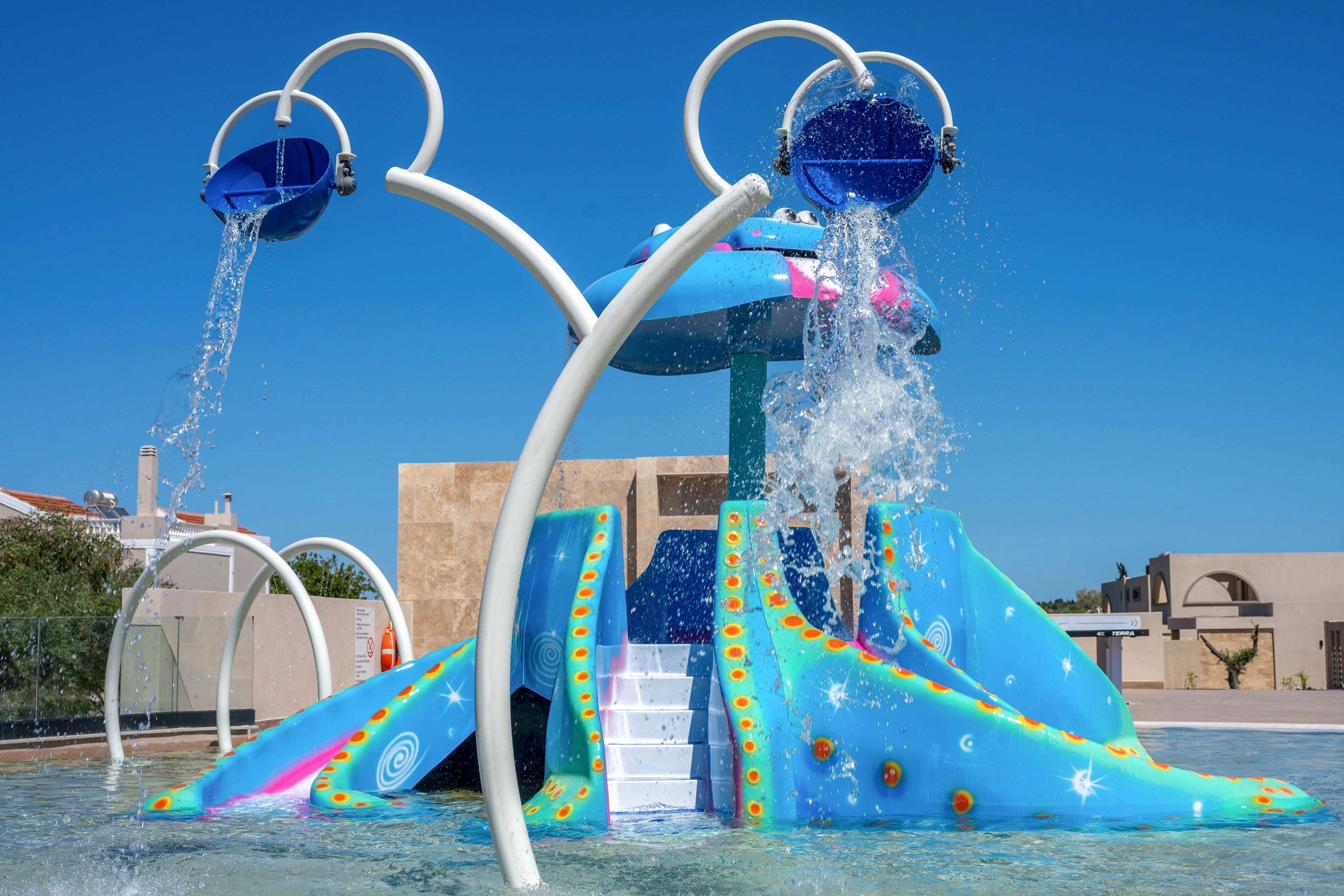 A bright blue aquatic playground structure stands in a shallow splash pad under a clear blue sky. Water pours from two overhead tipping buckets onto the slides and play area below. The structure features multiple slides, a central staircase, and whimsical painted patterns in orange, yellow, and white. Modern buildings and low walls are visible in the background, suggesting a resort or public recreation area.