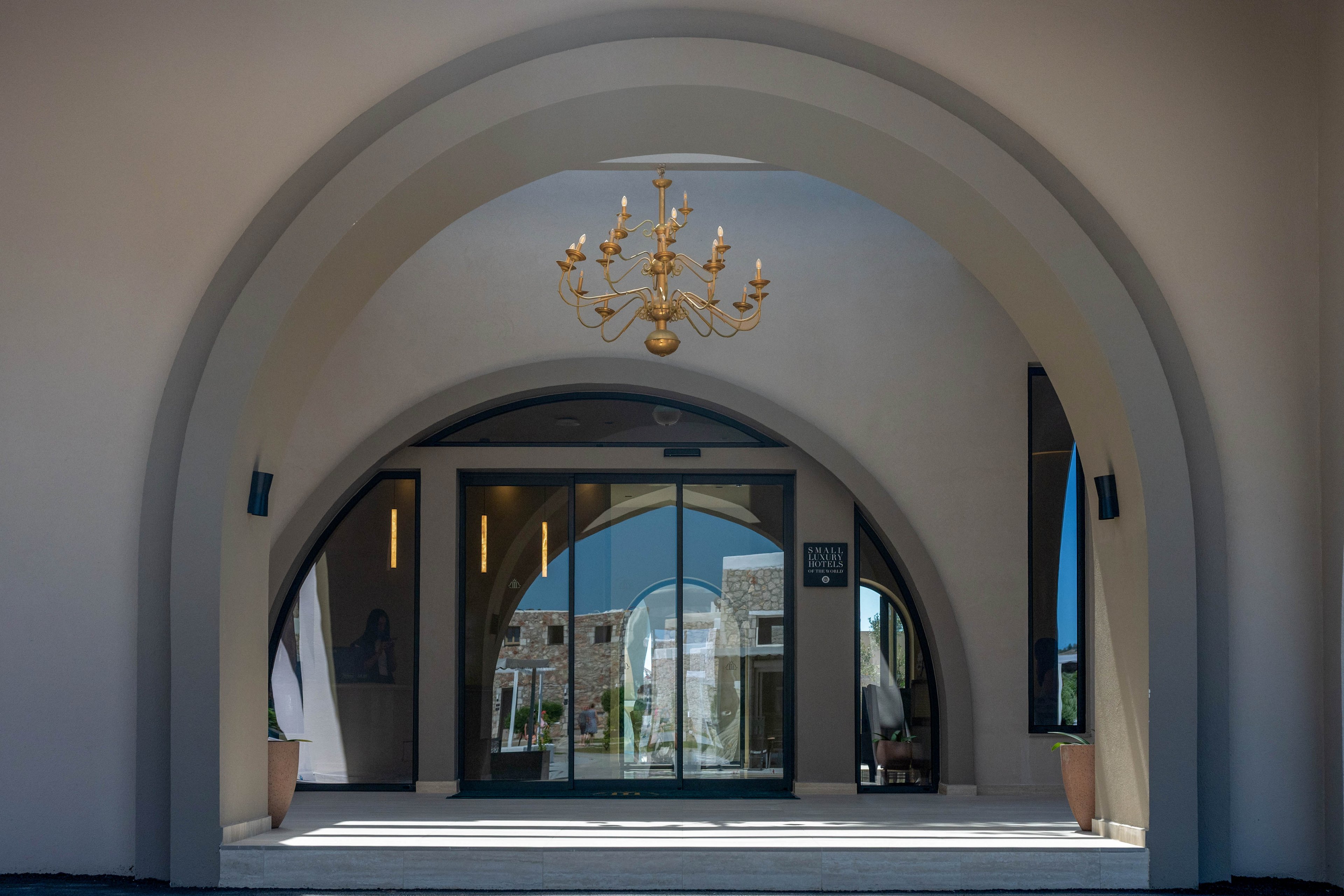 A grand entrance with multiple smooth arches frames a modern glass doorway. A gold chandelier hangs from the ceiling, adding a touch of luxury to the space. Through the glass doors, a courtyard and stone buildings are visible in the background. Two tall planters flank the entrance, enhancing the refined and welcoming atmosphere.