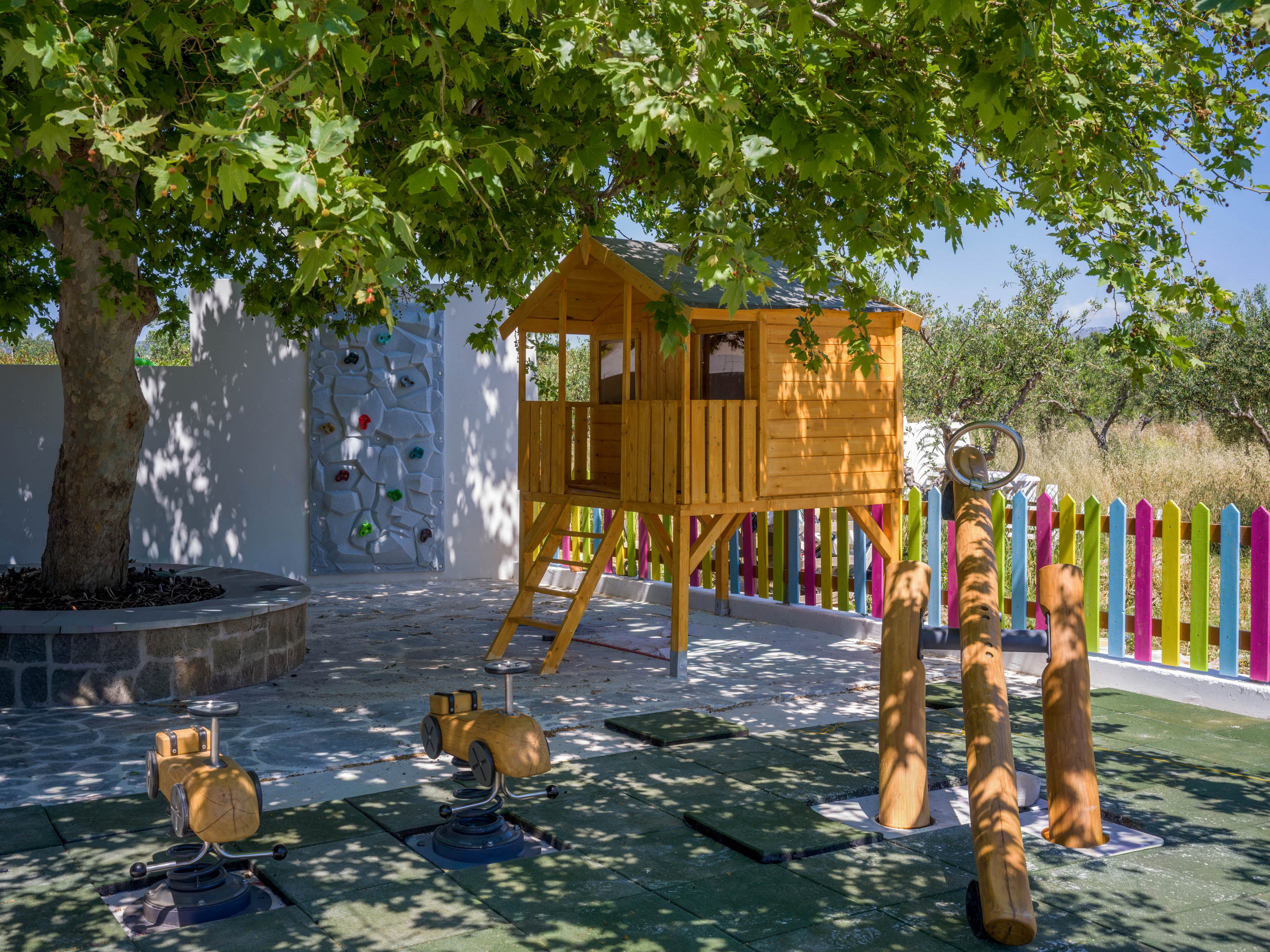 A wooden treehouse on stilts stands beneath a large leafy tree in a sunny outdoor playground. The play area includes spring-mounted animal rides and balance beams made of logs on a soft safety surface. A small climbing wall with colorful holds is built into a nearby wall. In the background, a multicolored picket fence separates the playground from a grassy field, creating a cheerful and inviting space for children.