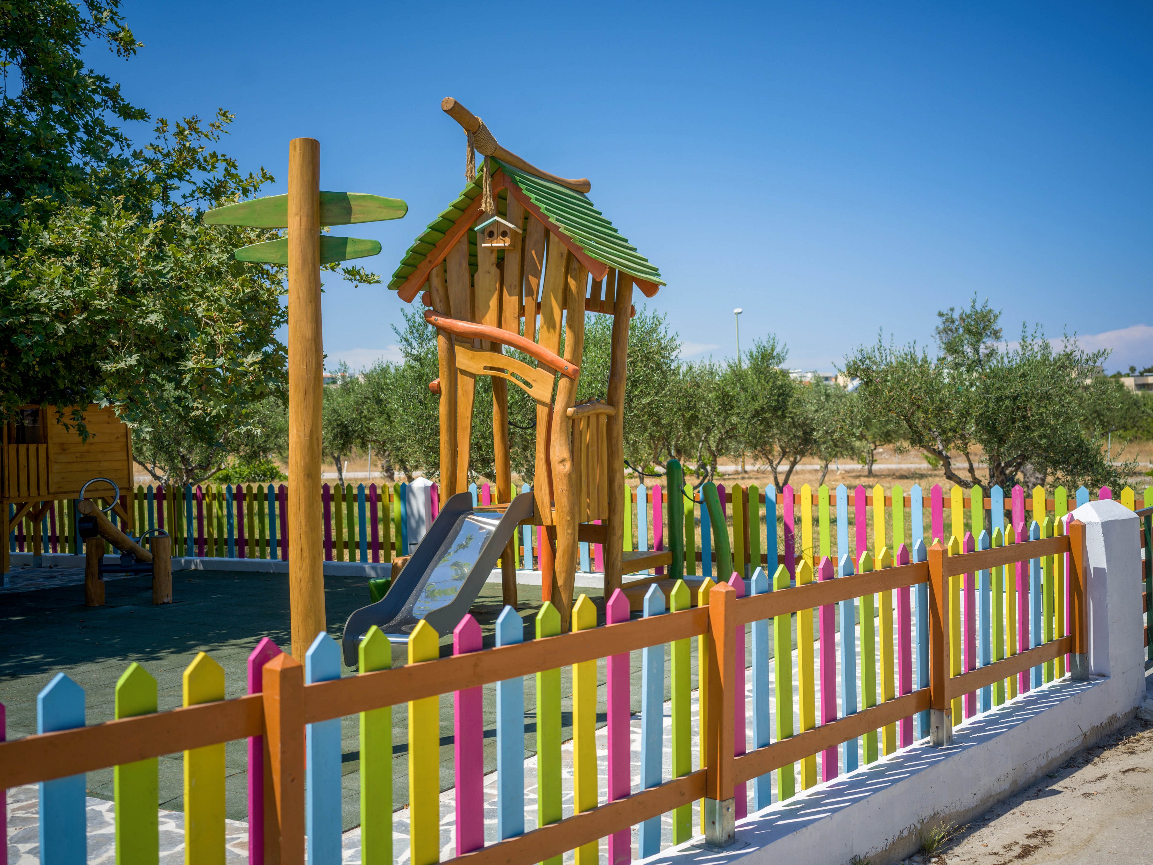 A small outdoor playground features a wooden play structure with a slide and climbing elements. The area is enclosed by a bright, multicolored picket fence that stands out against the green play surface. Trees and open fields surround the playground under a clear blue sky, suggesting a sunny day in a rural or suburban setting. The scene feels cheerful and inviting, ideal for young children.