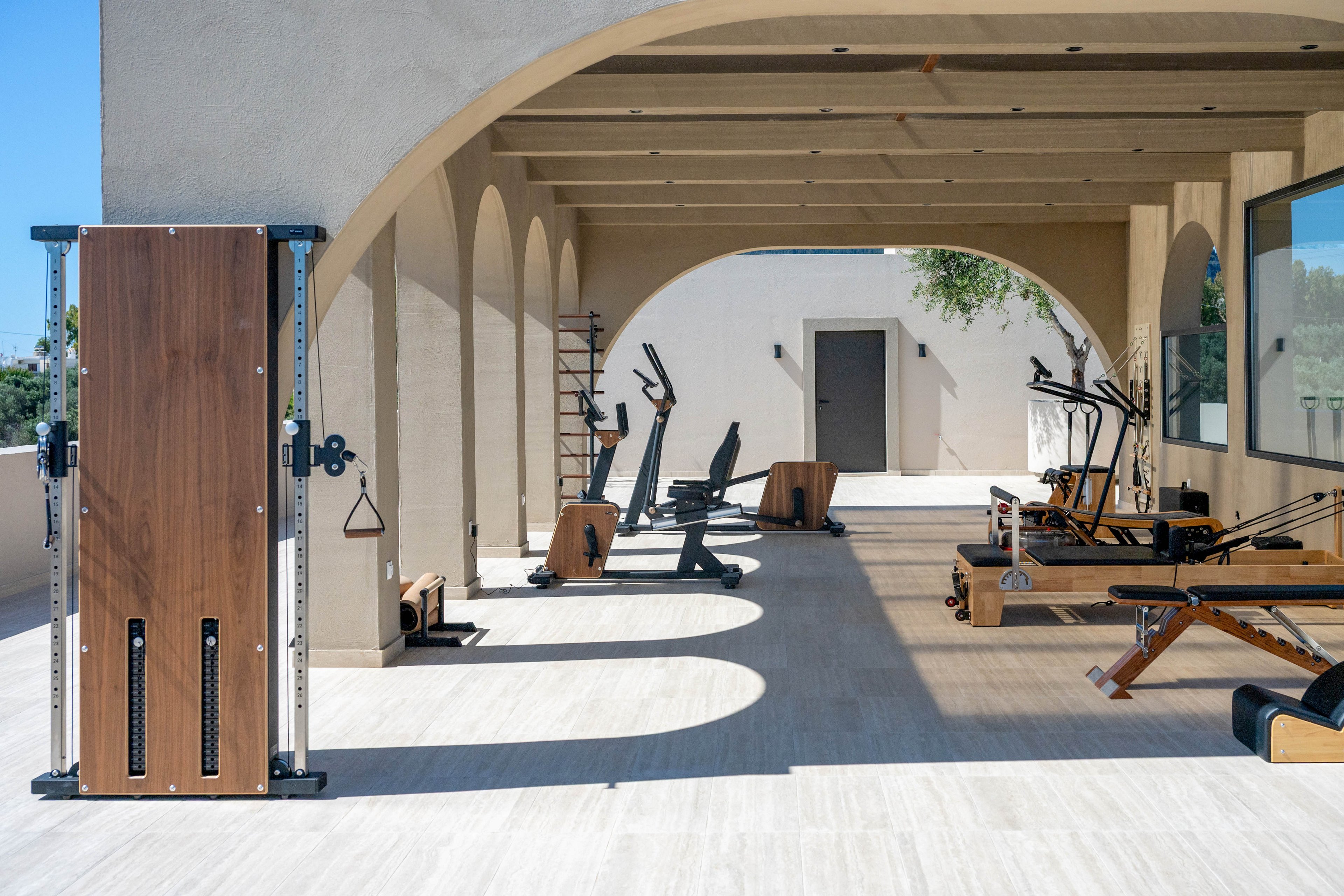 An open-air gym features a variety of modern exercise machines arranged under a covered terrace. Wooden and metal fitness equipment, including cardio and strength-training stations, lines the light-colored floor. Arched columns cast long shadows across the space, creating a pattern of light and shade. In the background, a closed door and minimal architectural details emphasize the clean, contemporary design.