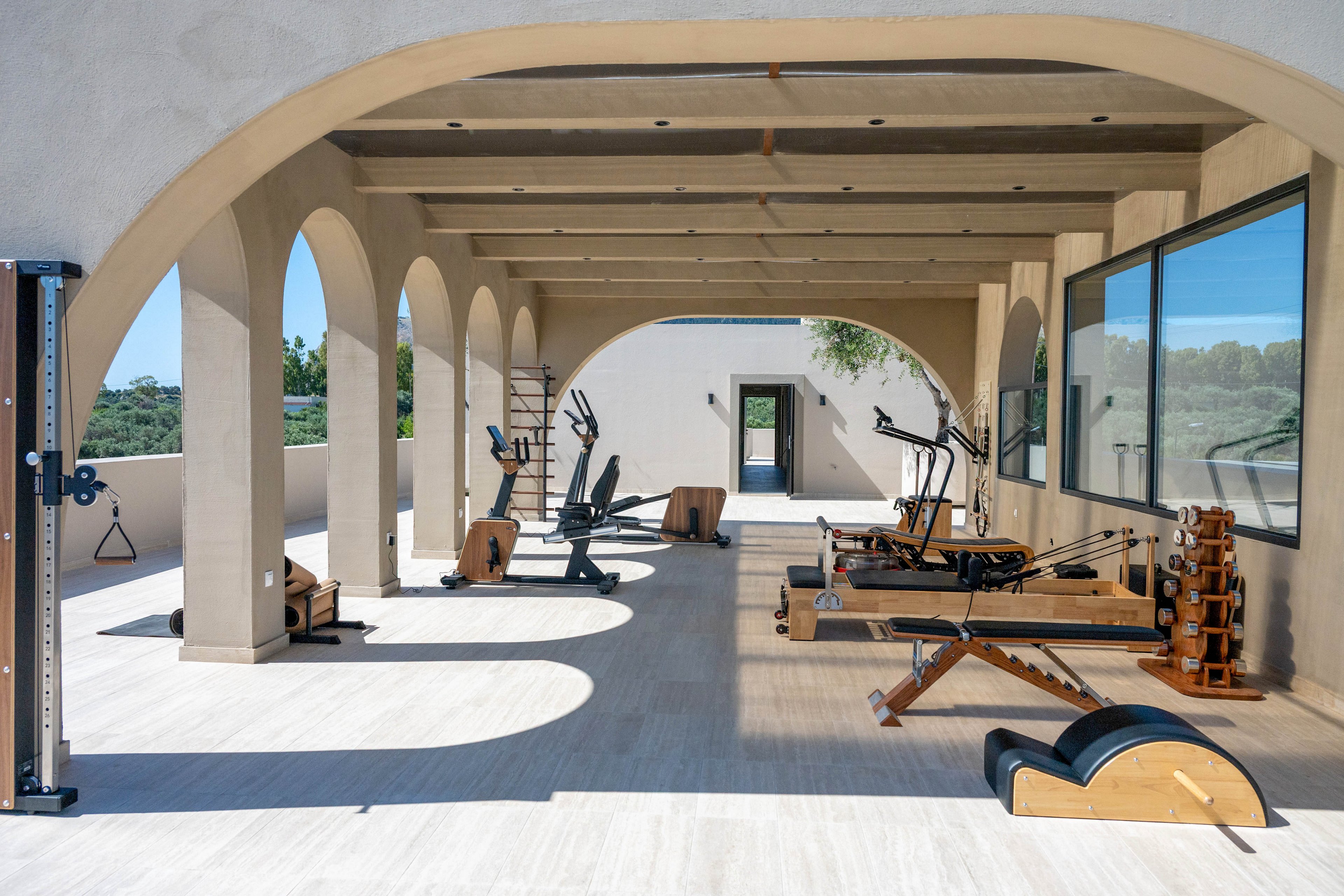 An outdoor covered gym features various wooden pilates and fitness machines arranged in a row. The space is set beneath an arched pergola attached to a modern home, with large windows on one side and open views on the other. Bright daylight casts strong shadows across the light-colored floor, emphasizing the clean architectural lines. The scene feels calm and luxurious, suggesting a private wellness or exercise area in a warm climate.