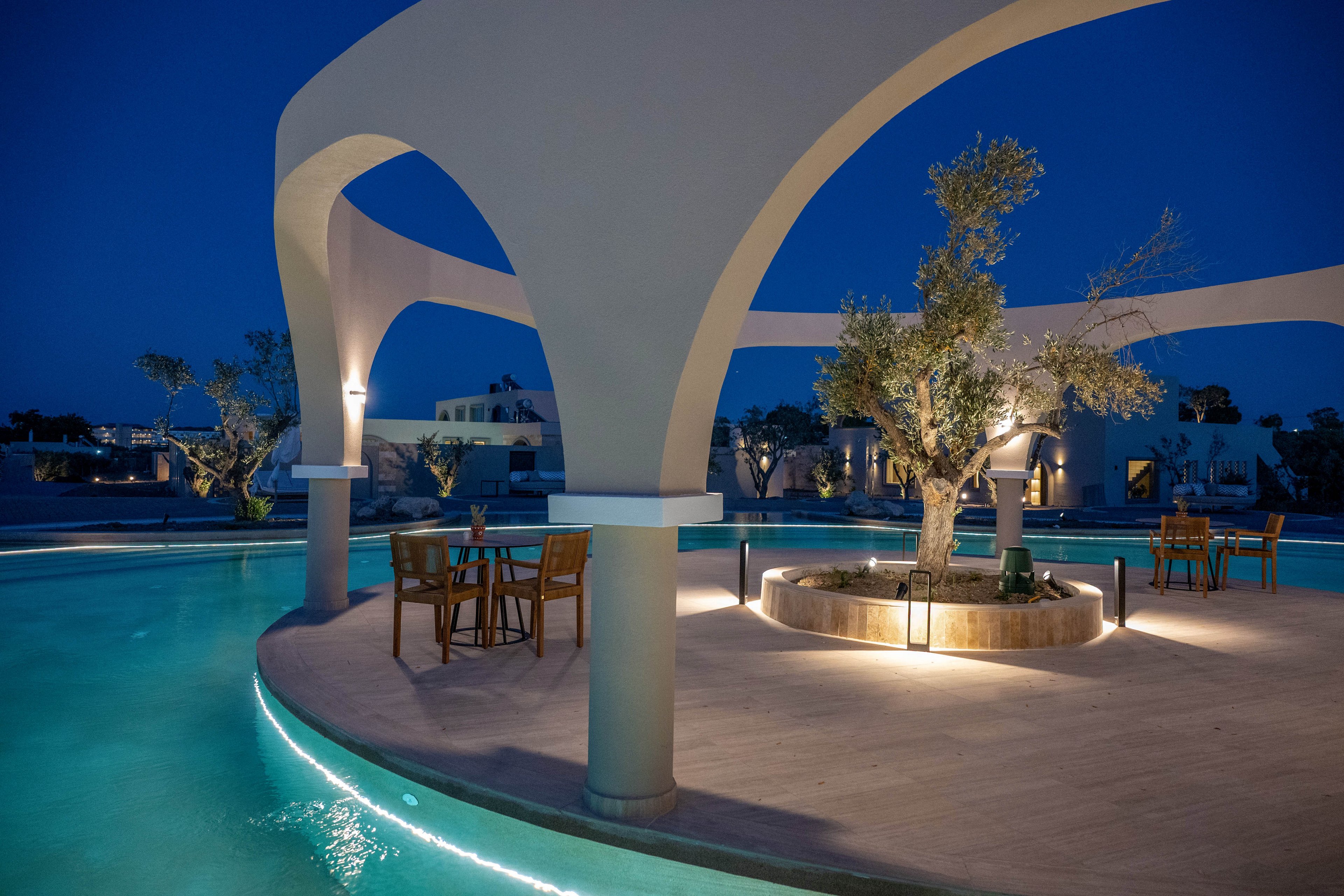 An elegant resort pool surrounds a circular terrace with a tree at its center, framed by smooth white arches. Soft lighting reflects on the turquoise water, creating a serene evening atmosphere. A few wooden tables and chairs are set up on the terrace for outdoor dining or relaxation. Modern buildings and additional trees are visible in the background under a deep blue twilight sky.