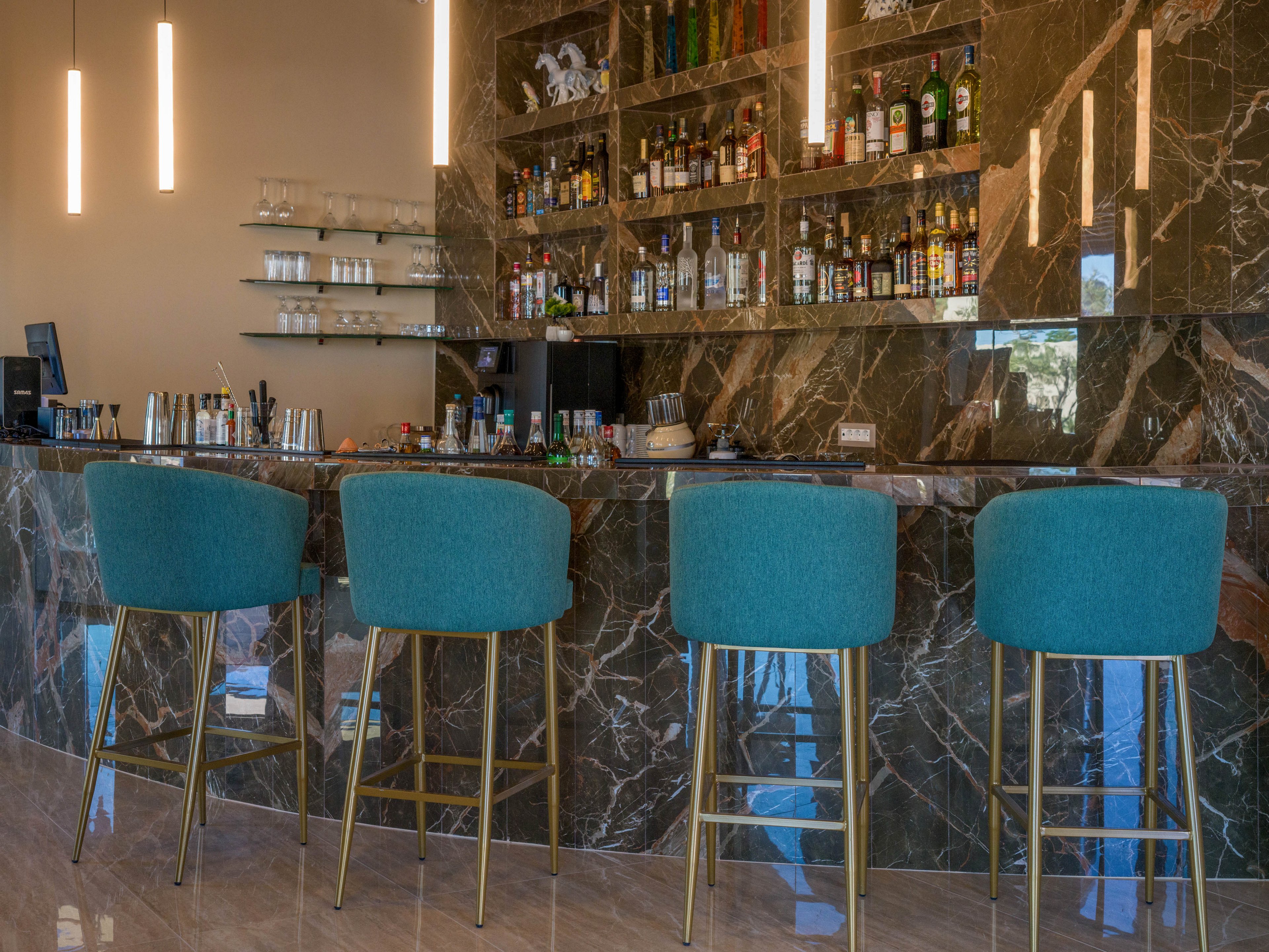 A contemporary bar interior features a polished dark marble counter and matching wall. Four teal upholstered bar stools with metal legs line the front of the bar. Shelves behind the counter display an assortment of liquor bottles and glassware. Minimalist pendant lights hang from the ceiling, creating a warm and inviting atmosphere.
