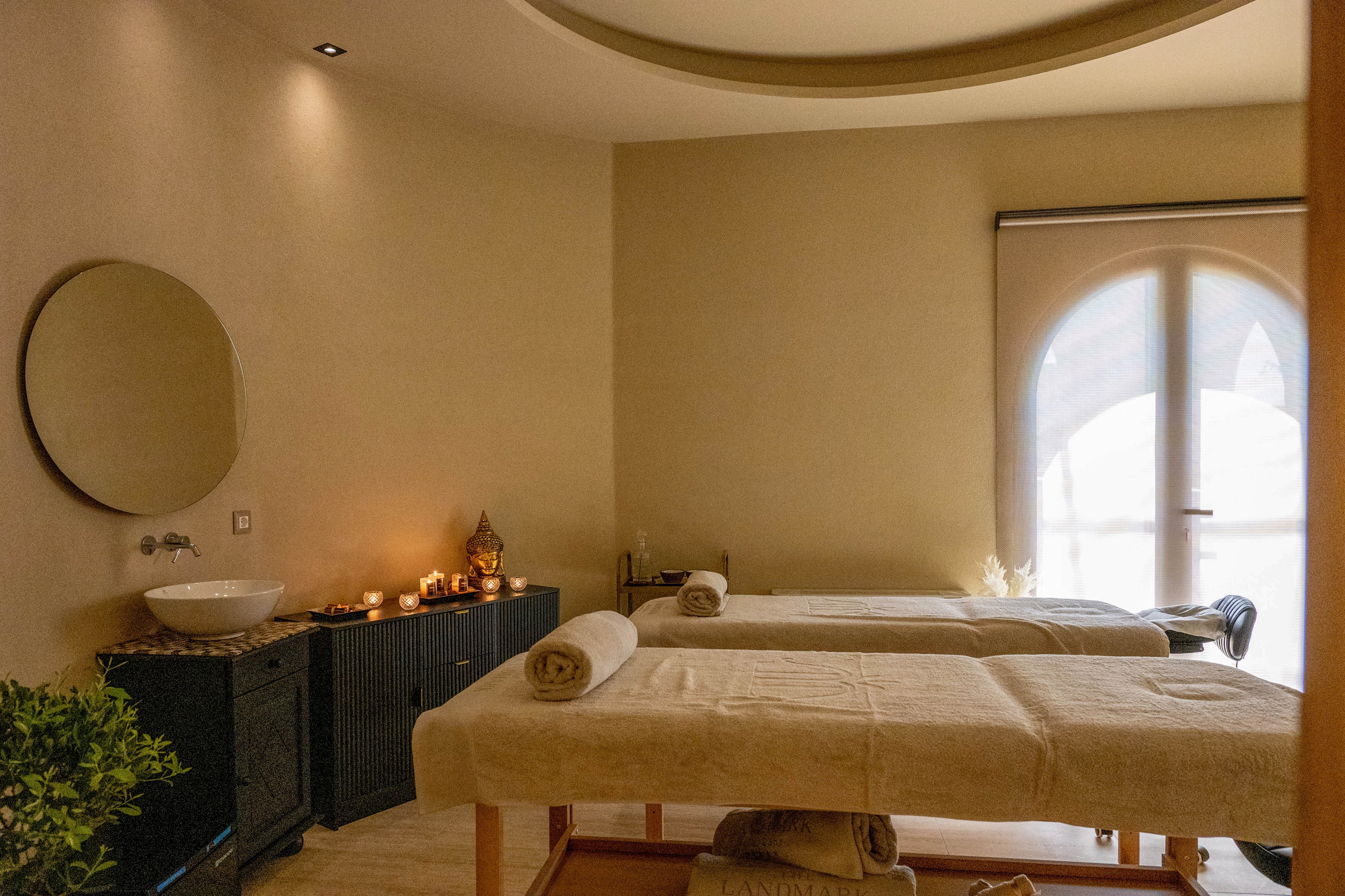 A warm, softly lit spa treatment room features two massage tables neatly prepared with rolled towels. A side cabinet holds a white basin and several lit candles, adding a relaxing glow to the space. Neutral beige walls and minimal decor create a calm, uncluttered atmosphere. Natural light filters through a large arched window with a shade, enhancing the serene and inviting mood.
