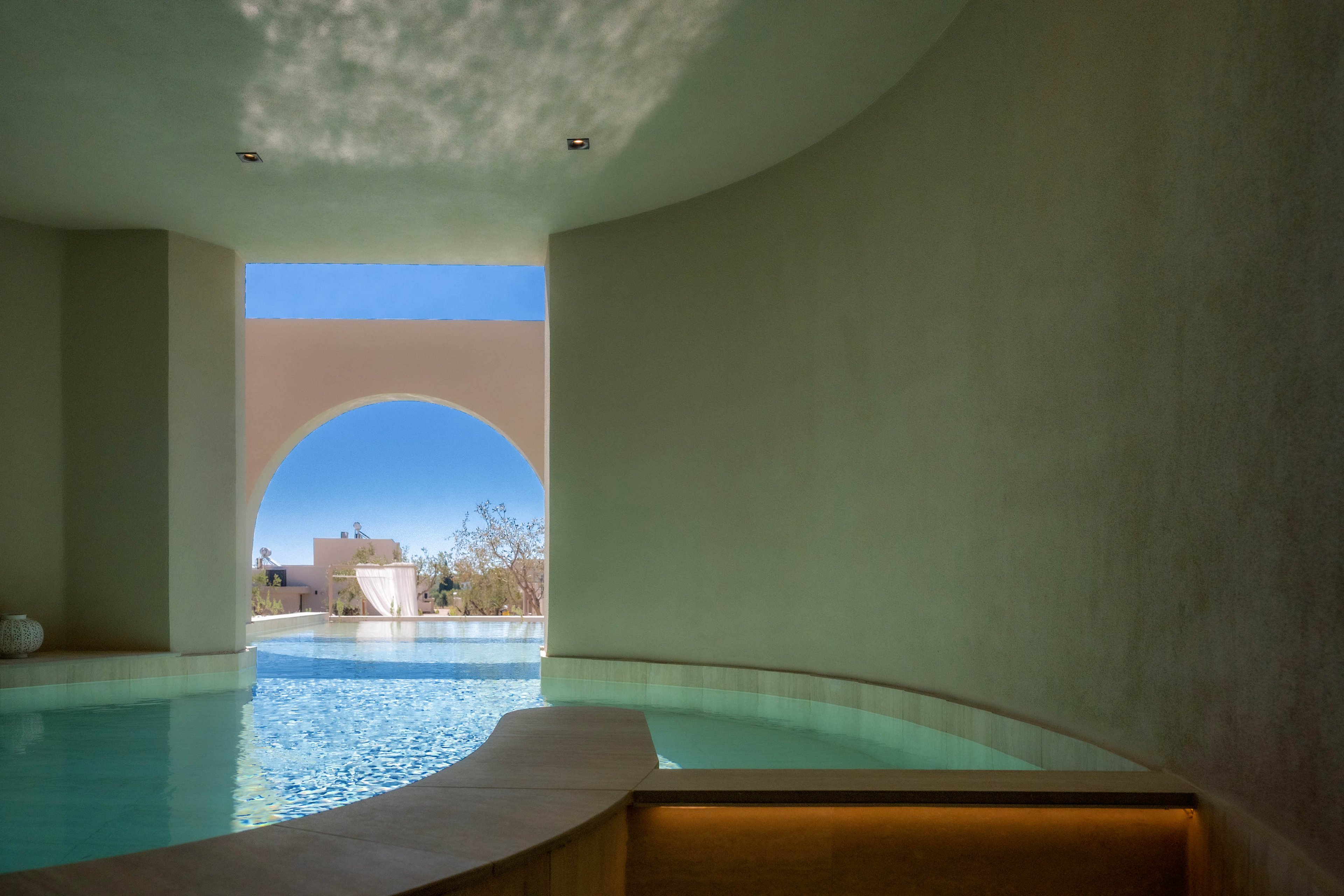 A serene indoor pool curves along smooth, minimalist walls. Natural light pours in through a large exterior archway framing a bright blue sky and distant buildings. The water reflects soft shadows on the ceiling, enhancing the tranquil atmosphere. Warm recessed lighting under a bench contrasts with the cool aqua tones of the pool.