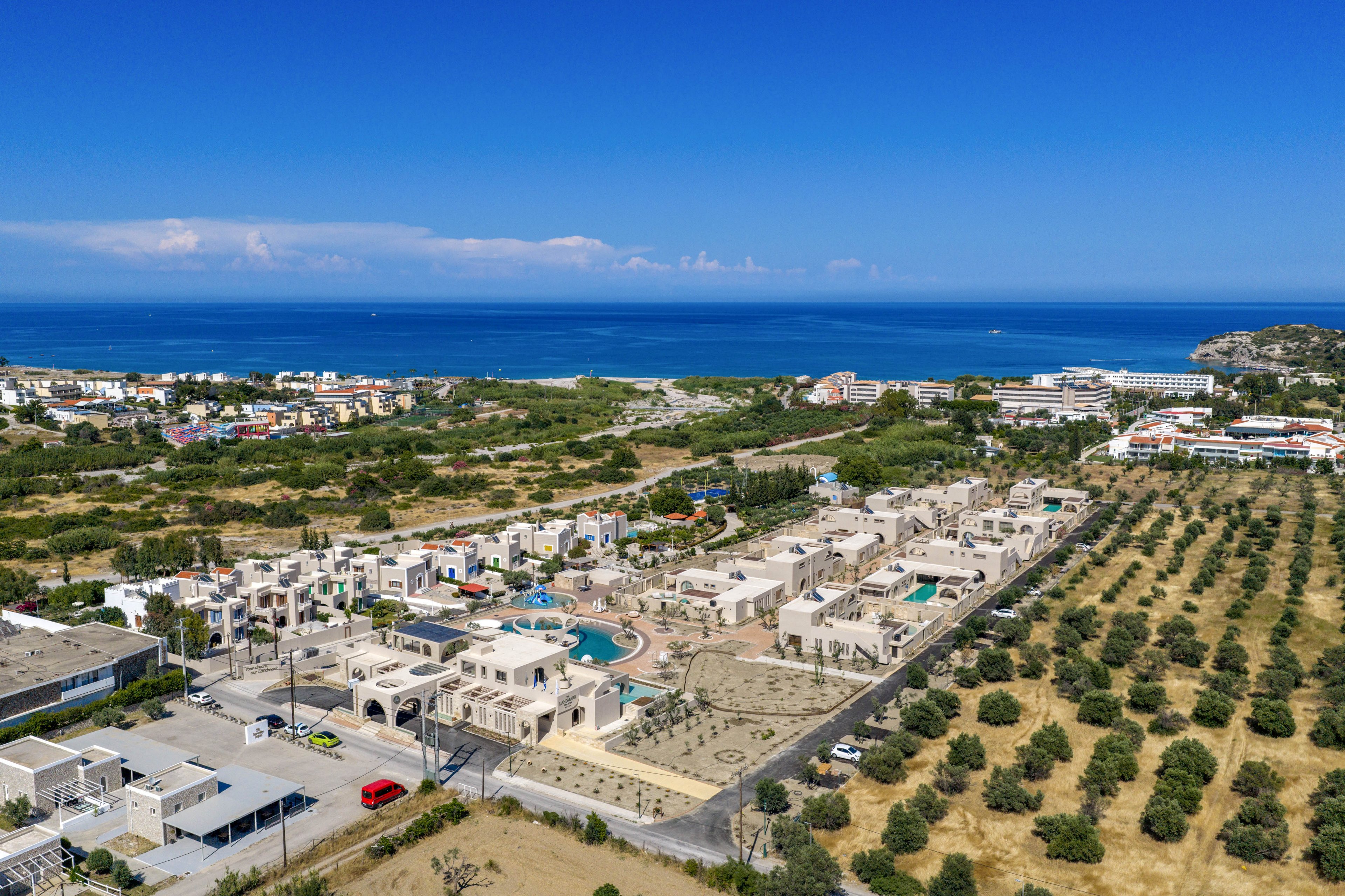 A coastal resort complex with white low-rise buildings and swimming pools is captured from an aerial perspective. The property is surrounded by open fields and orderly rows of trees, likely an agricultural or orchard area. In the distance, additional buildings and small settlements stretch toward the shoreline. The deep blue sea and clear sky create a bright, summery atmosphere. Roads, parked cars, and landscaped areas add detail to this Mediterranean-style landscape.