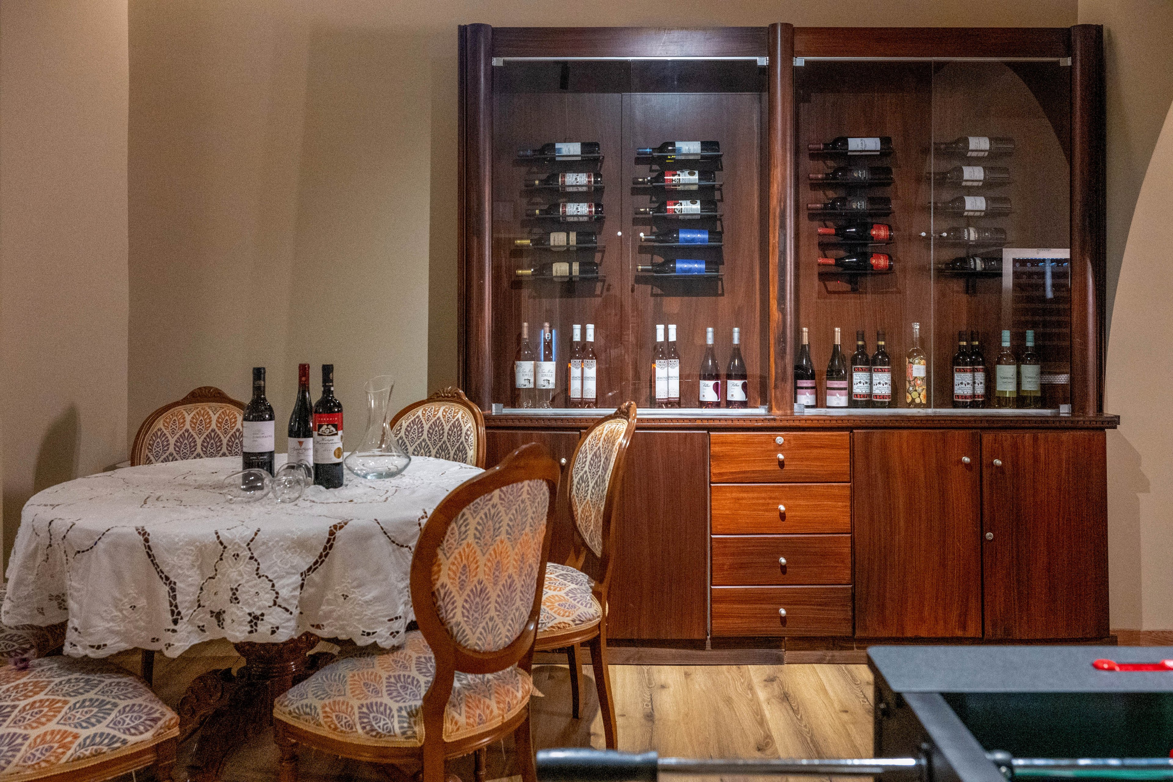 A cozy wine room features a round table covered with a lace tablecloth and surrounded by upholstered wooden chairs. Several bottles of wine and empty wine glasses are arranged on the table, ready for tasting. Behind the seating area, a large wooden cabinet displays numerous wine bottles on shelves and in glass-fronted sections. Warm lighting and wooden flooring create an inviting, intimate atmosphere suitable for entertaining.