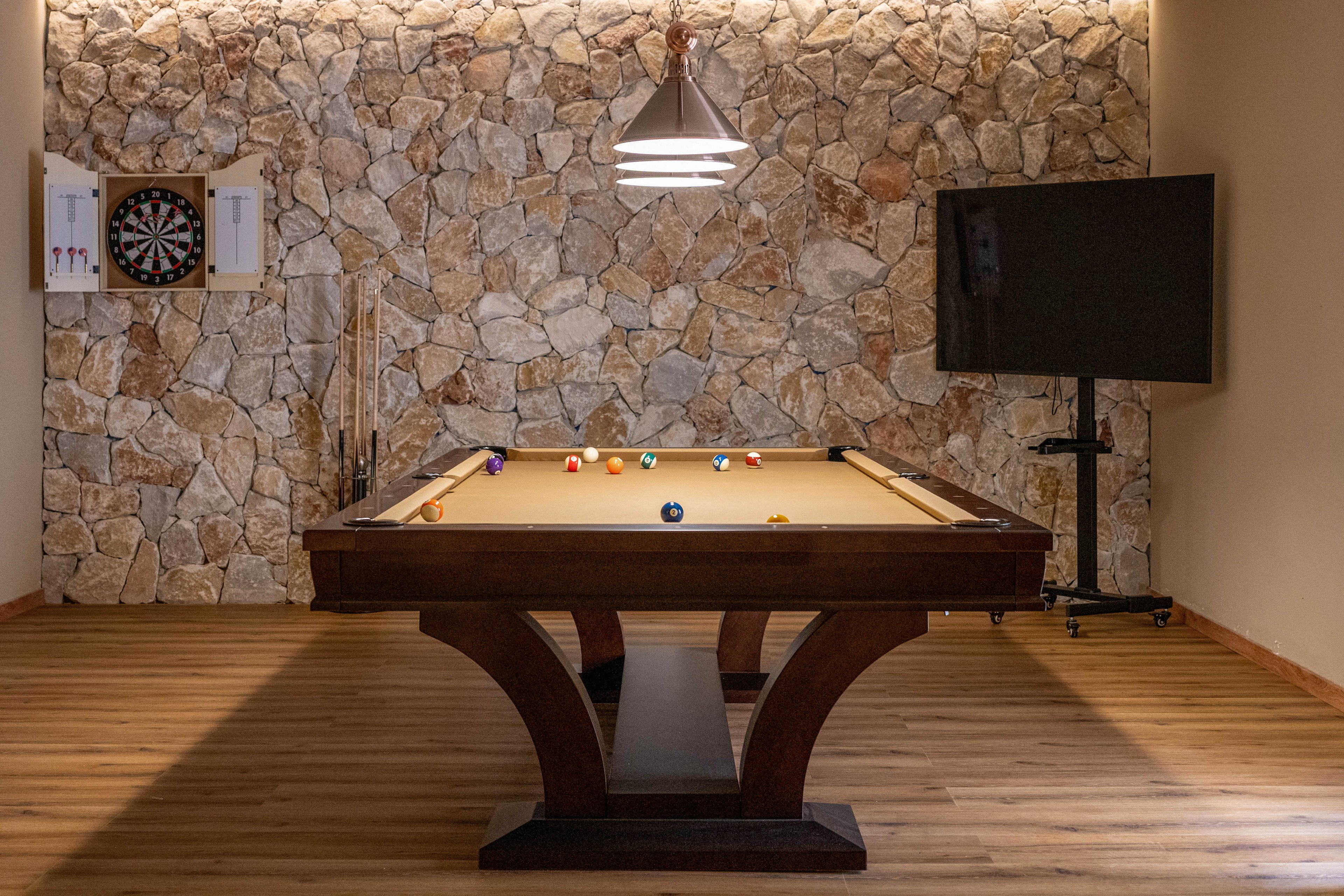 A contemporary billiards table stands centered in a cozy game room. Pool balls are scattered across the felt surface under a hanging pendant light. The room features a stone accent wall, wooden flooring, and a mounted dartboard on the left. A large flat-screen television on a stand is positioned to the right, completing the entertainment space.