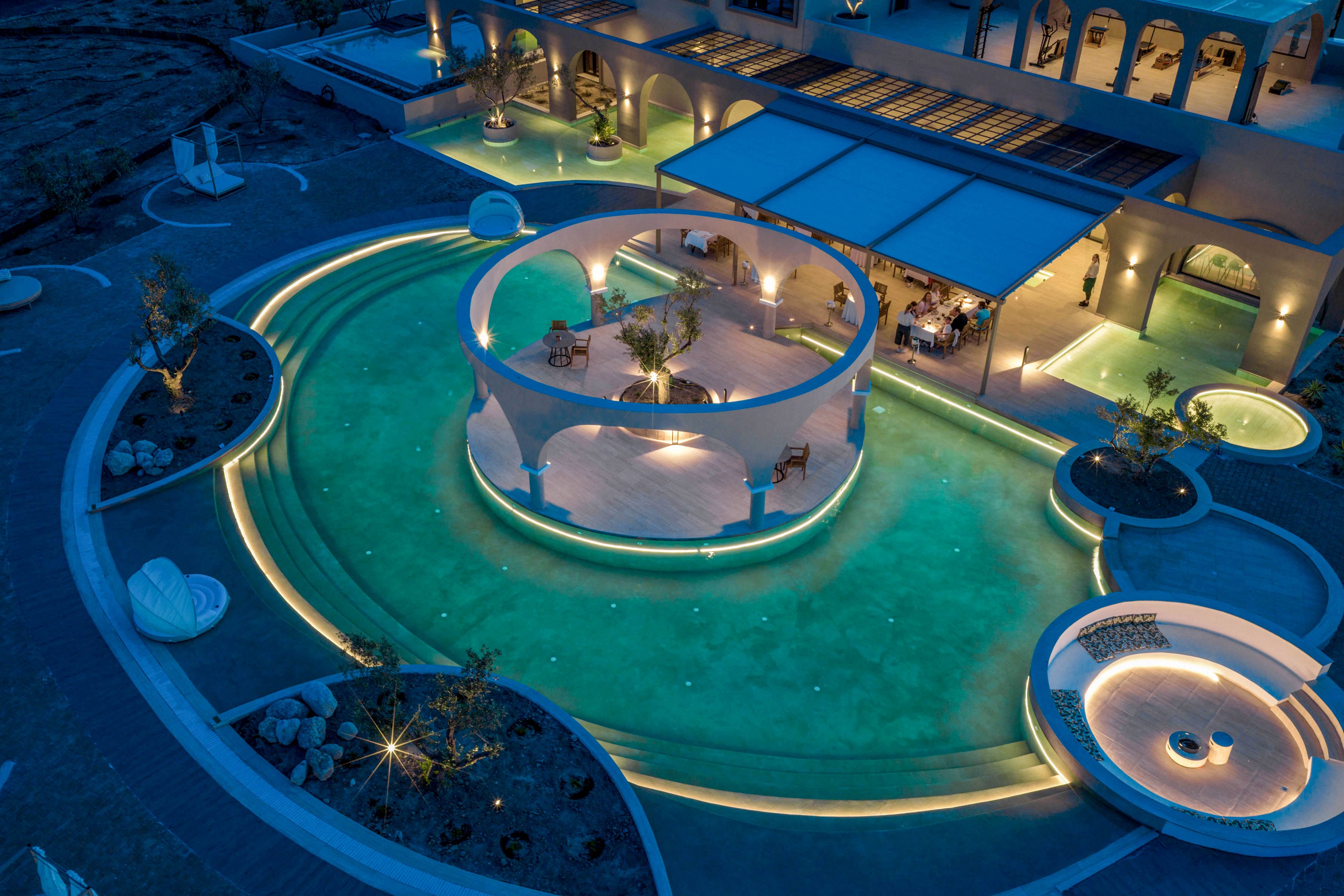 A luxurious resort features an illuminated pool complex captured at night. Curved walkways and circular islands create an elegant, sculptural layout within the water. Soft blue and warm yellow lighting highlight the architecture and outdoor seating areas. The surrounding building with arches and terraces adds to the upscale, tranquil atmosphere.