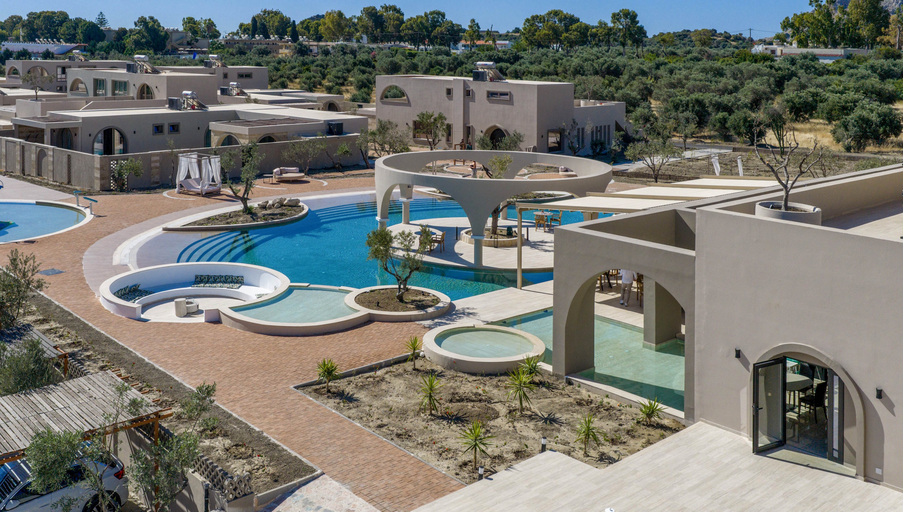 A contemporary resort complex features multiple curved swimming pools surrounded by beige, cube-like buildings. The architecture includes open-air arches, circular islands, and shaded lounge areas integrated into the pool design. Pathways of warm-toned stone connect the structures, with young plants and trees adding greenery to the courtyard. In the background, additional villas and dense trees create a secluded, tranquil vacation setting.
