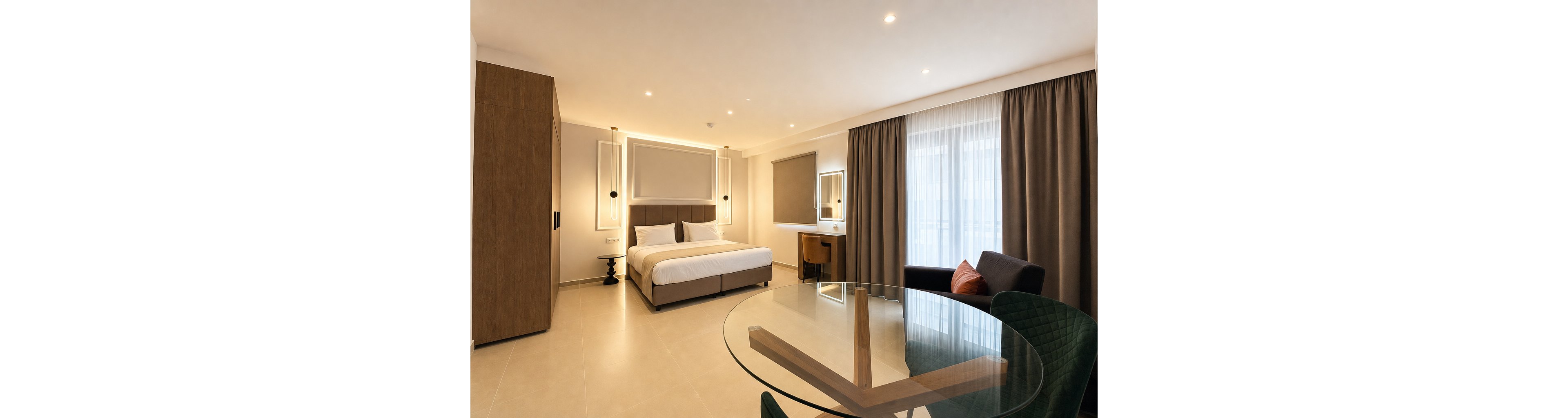 A spacious modern bedroom features a large bed with white linens and a padded headboard. The room includes built-in wooden wardrobes, a small desk with a chair, and contemporary wall lighting. Floor-to-ceiling curtains cover a wide window or balcony door, allowing soft natural light into the space. In the foreground, a round glass table and upholstered chairs add a touch of elegance to the minimalist interior.