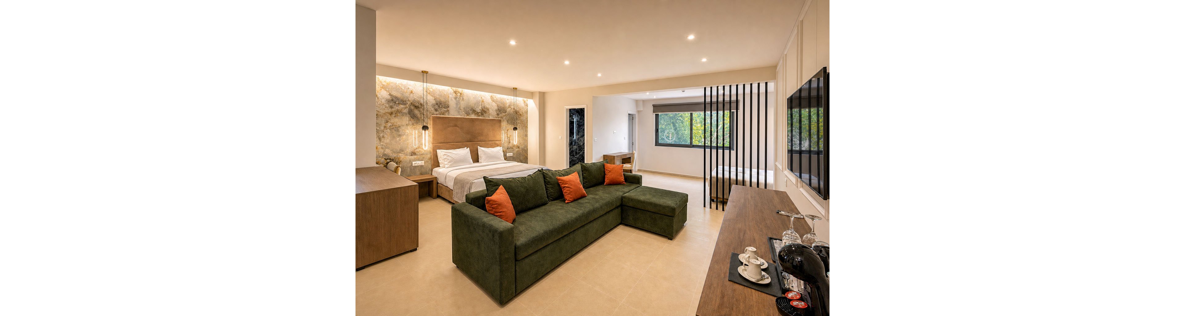 A spacious modern hotel suite features a large bed with white linens and a warm-toned headboard against a textured accent wall. In the foreground, a green sectional sofa with orange cushions faces a wooden console and wall-mounted television. The room includes a work desk, a coffee and tea station with cups and kettle, and a partially open area separated by vertical black slats. Large windows bring in natural light and offer a view of greenery outside, enhancing the calm and inviting atmosphere.