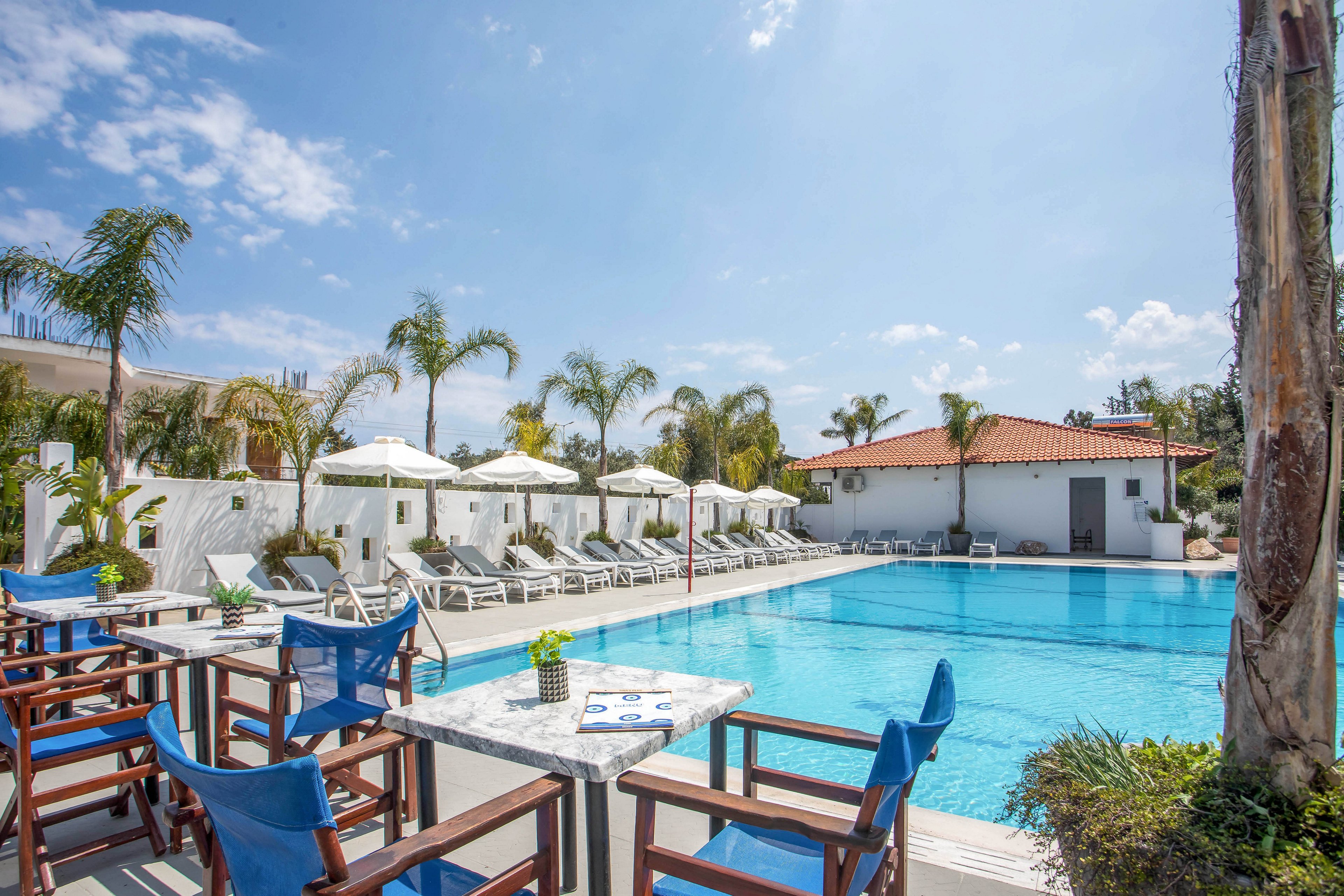 An outdoor resort swimming pool is surrounded by white lounge chairs and umbrellas under a bright blue sky. Wooden chairs with blue cushions and marble-topped tables sit in the foreground near the pool edge. Palm trees and white walls frame the scene, with a tiled-roof building in the background. The atmosphere is calm and inviting, suggesting a relaxing vacation setting.