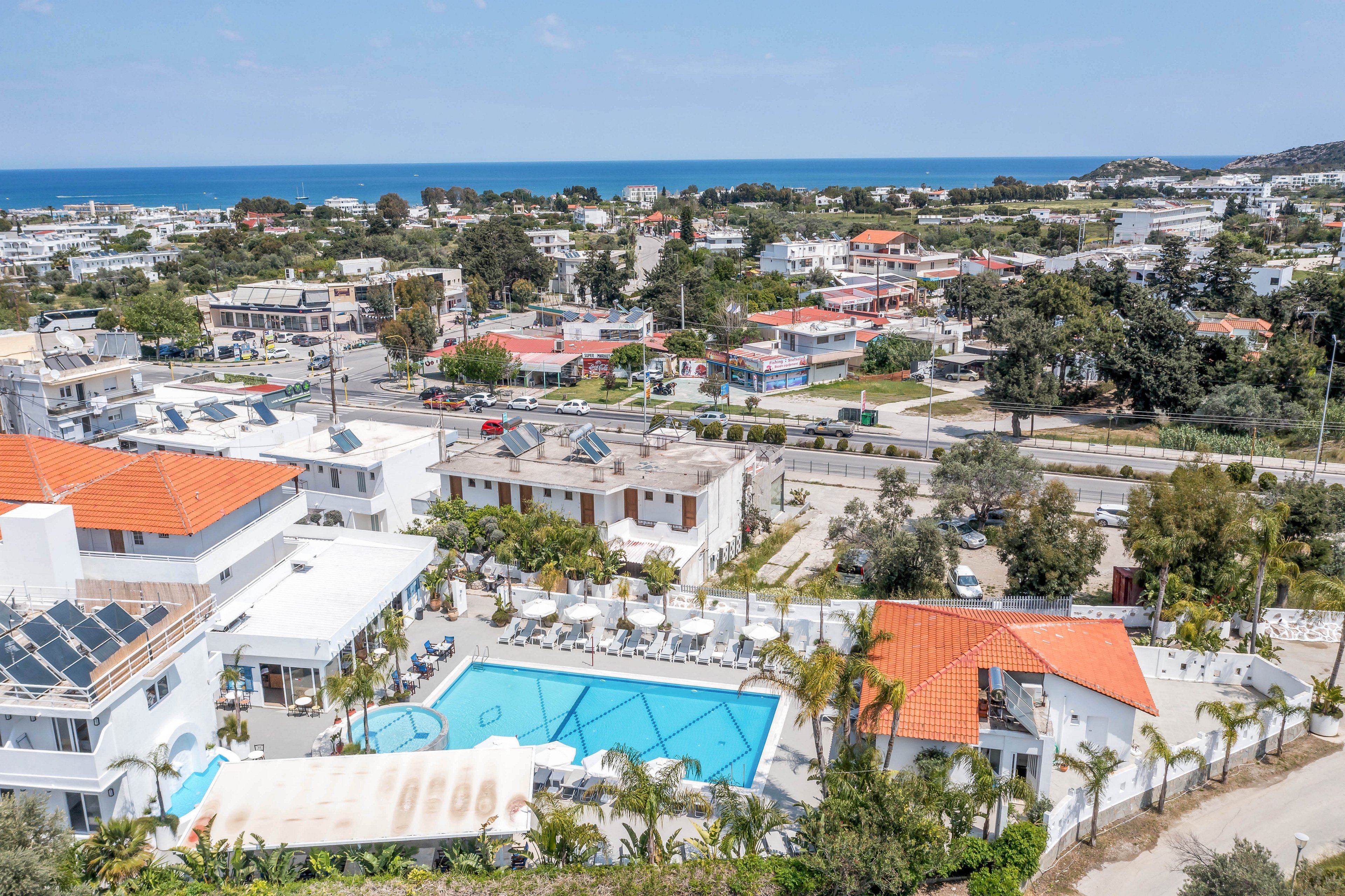 A coastal resort complex with a large outdoor swimming pool is captured from an elevated aerial perspective. White buildings with red-tiled roofs surround the pool area, which is lined with sun loungers and umbrellas. The resort is set within a small town featuring low-rise houses, roads, and scattered greenery. In the distance, the blue sea and coastline stretch across the horizon under a clear sky.