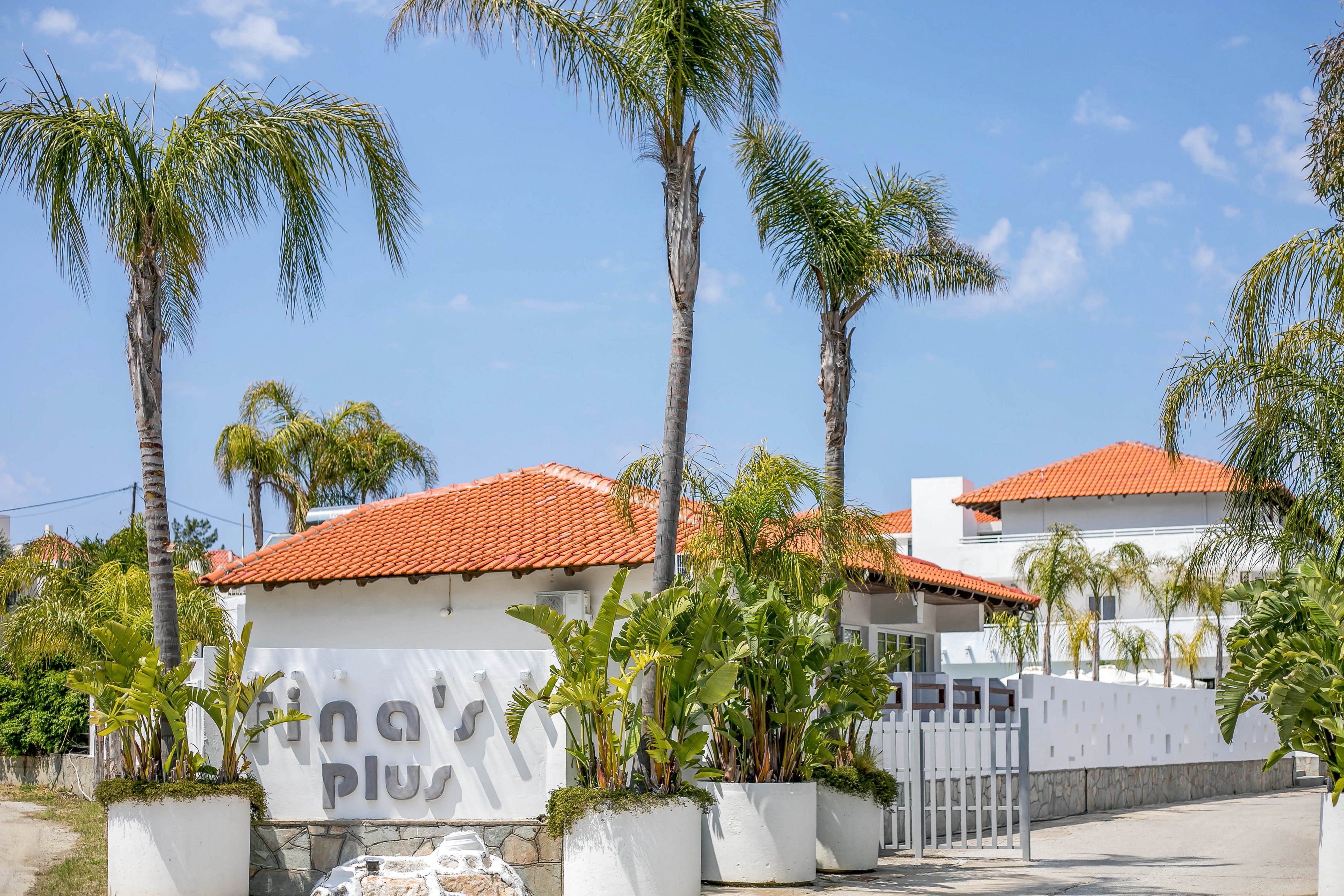 A white-walled villa complex with red tile roofs is surrounded by tall palm trees under a bright blue sky. The entrance area features large white planters filled with lush green plants and a metal gate. Text is partially visible on the front wall, suggesting the name of the property, though it is not fully legible. The overall scene evokes a sunny, Mediterranean or tropical resort atmosphere.