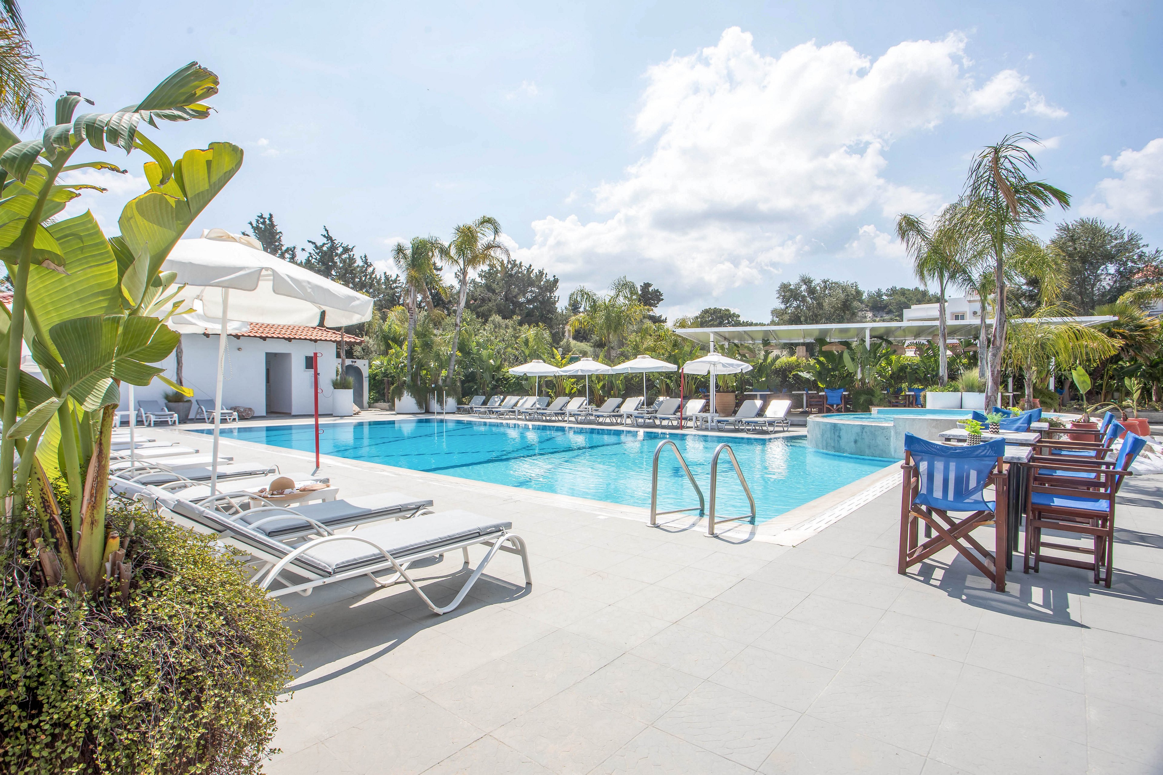 An outdoor resort swimming pool is surrounded by white lounge chairs and umbrellas on a bright sunny day. The pool area is framed by tropical plants and palm trees, creating a lush, inviting atmosphere. A row of tables and chairs sits to the side, suggesting a nearby bar or dining area. Clear blue water, white architecture, and open sky emphasize a clean, relaxing vacation setting.