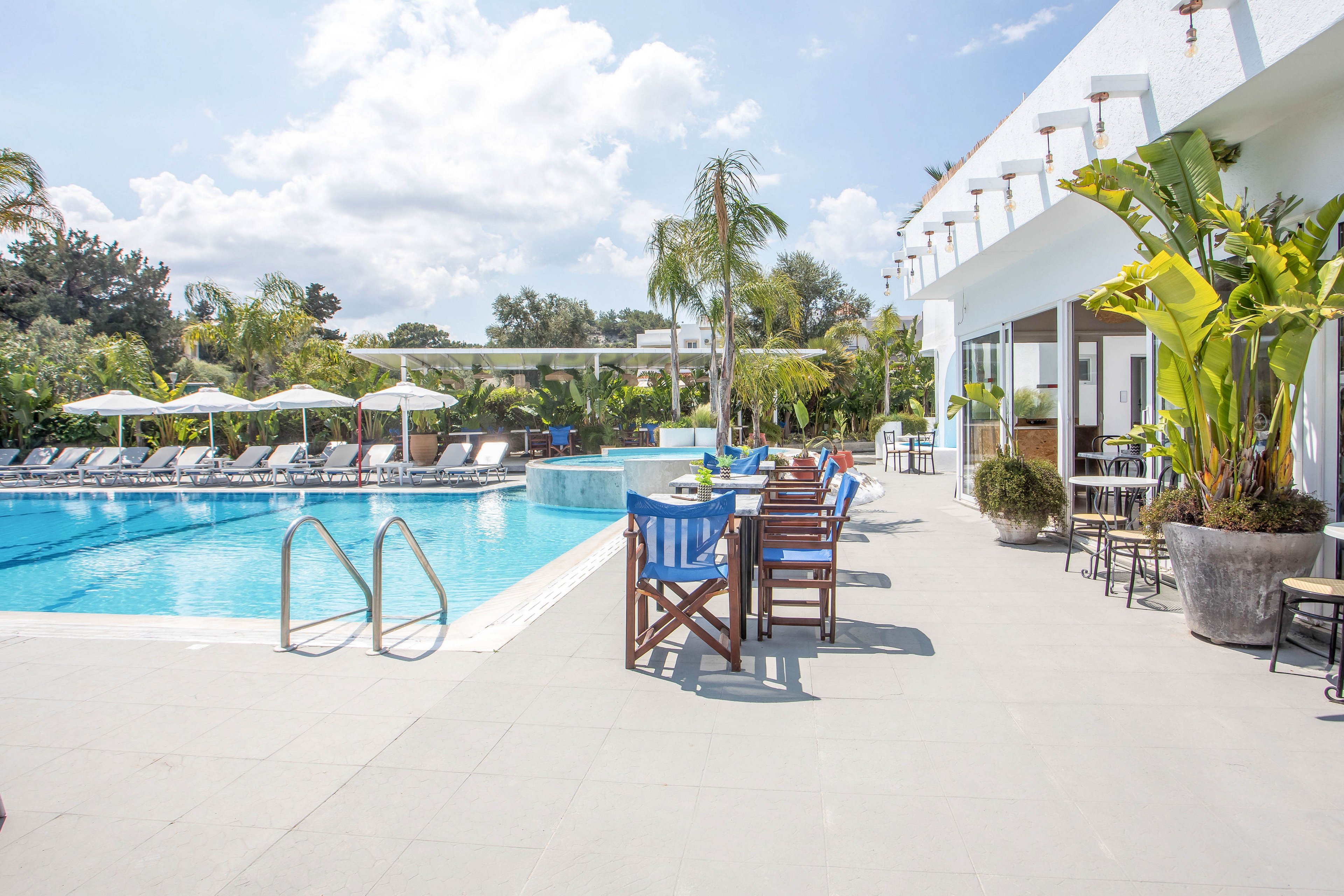 A bright outdoor swimming pool is surrounded by lounge chairs and umbrellas at a modern resort. Wooden tables and chairs line the poolside terrace next to a white building with large windows. Tropical plants and palm trees add lush greenery around the water. The clear sky and sunlight create a relaxed vacation atmosphere.