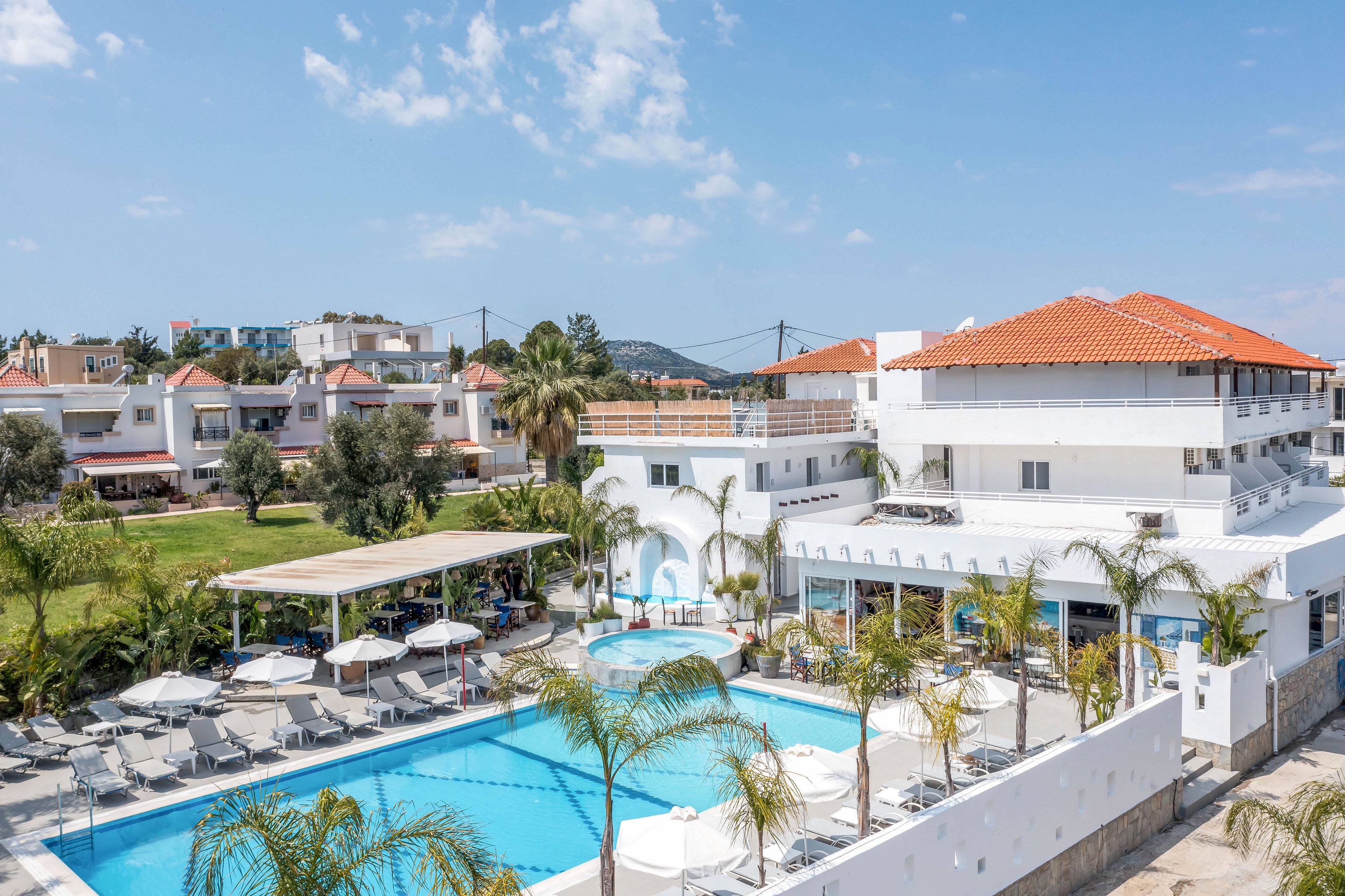 A bright resort hotel with white buildings and red-tiled roofs surrounds a large outdoor swimming pool. Sun loungers and umbrellas line the poolside terrace, offering shaded relaxation areas. Palm trees and green lawns add a tropical touch to the scene, while neighboring houses are visible in the background. The clear blue sky and calm atmosphere suggest a warm, sunny vacation destination.