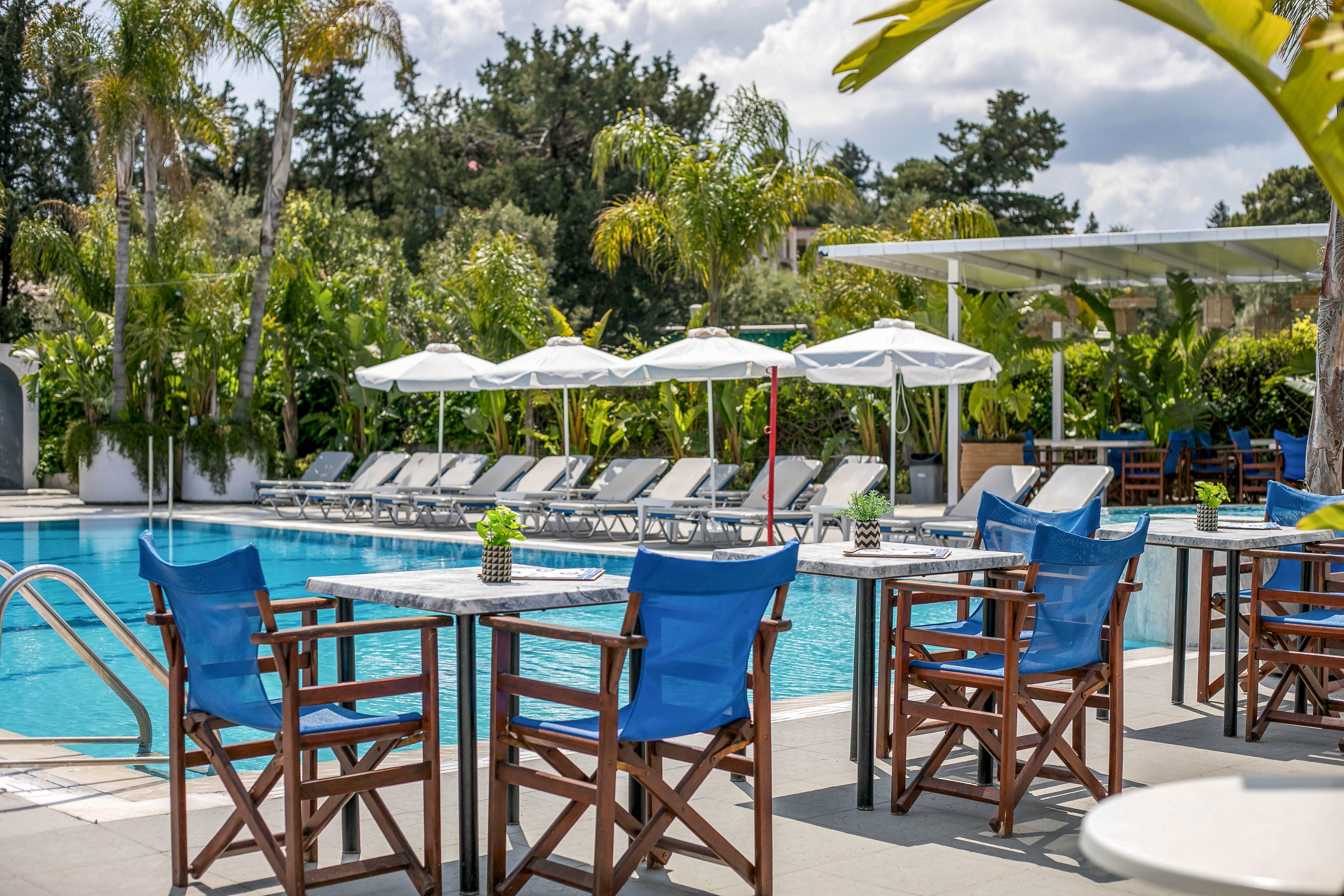 An inviting outdoor swimming pool is surrounded by lounge chairs and white umbrellas at a resort. Wooden tables with blue canvas chairs are arranged in the foreground, suggesting a casual poolside bar or café area. Lush green palm trees and tropical plants frame the scene, creating a private oasis atmosphere. The bright daylight and clear water convey a relaxing vacation mood.