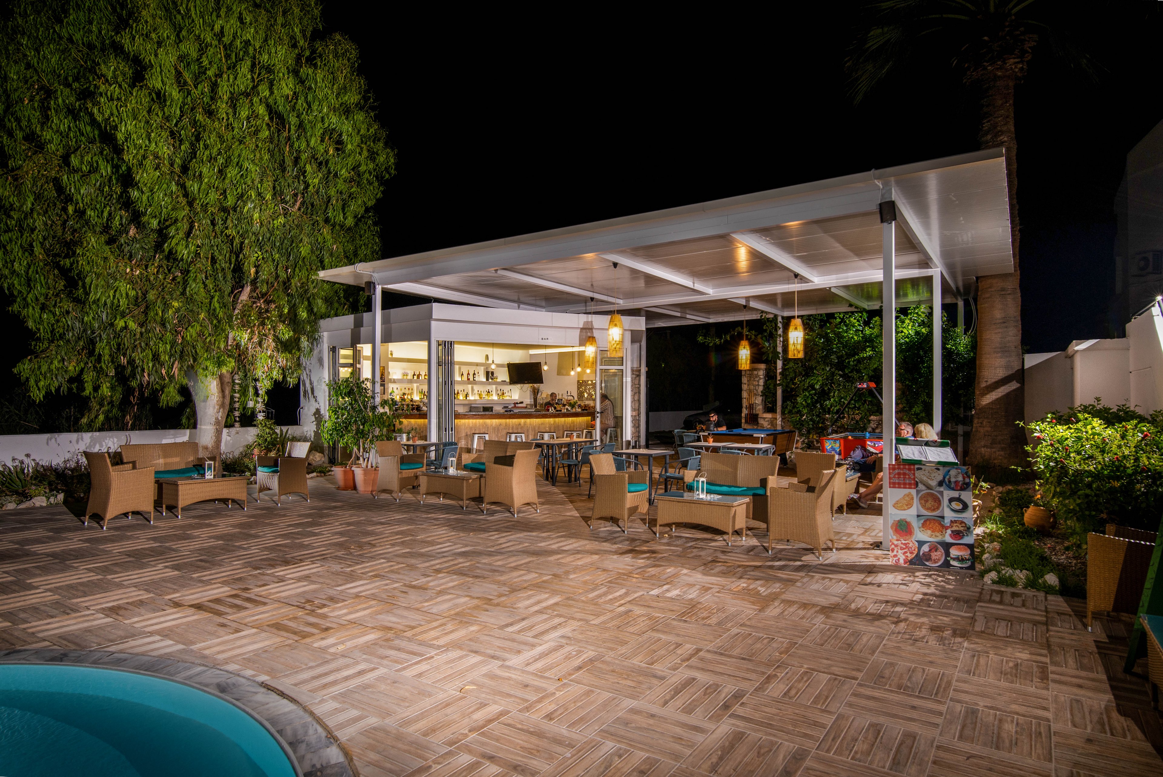 A contemporary outdoor restaurant patio is shown at night, featuring a modern open-air bar with shelves of bottles and glassware. The seating area includes wicker chairs and tables arranged under a white canopy, with warm lighting creating a cozy ambiance. Lush greenery surrounds the space, and a pool edge is visible in the foreground. The scene is quiet, with no people present, emphasizing the inviting nighttime atmosphere.