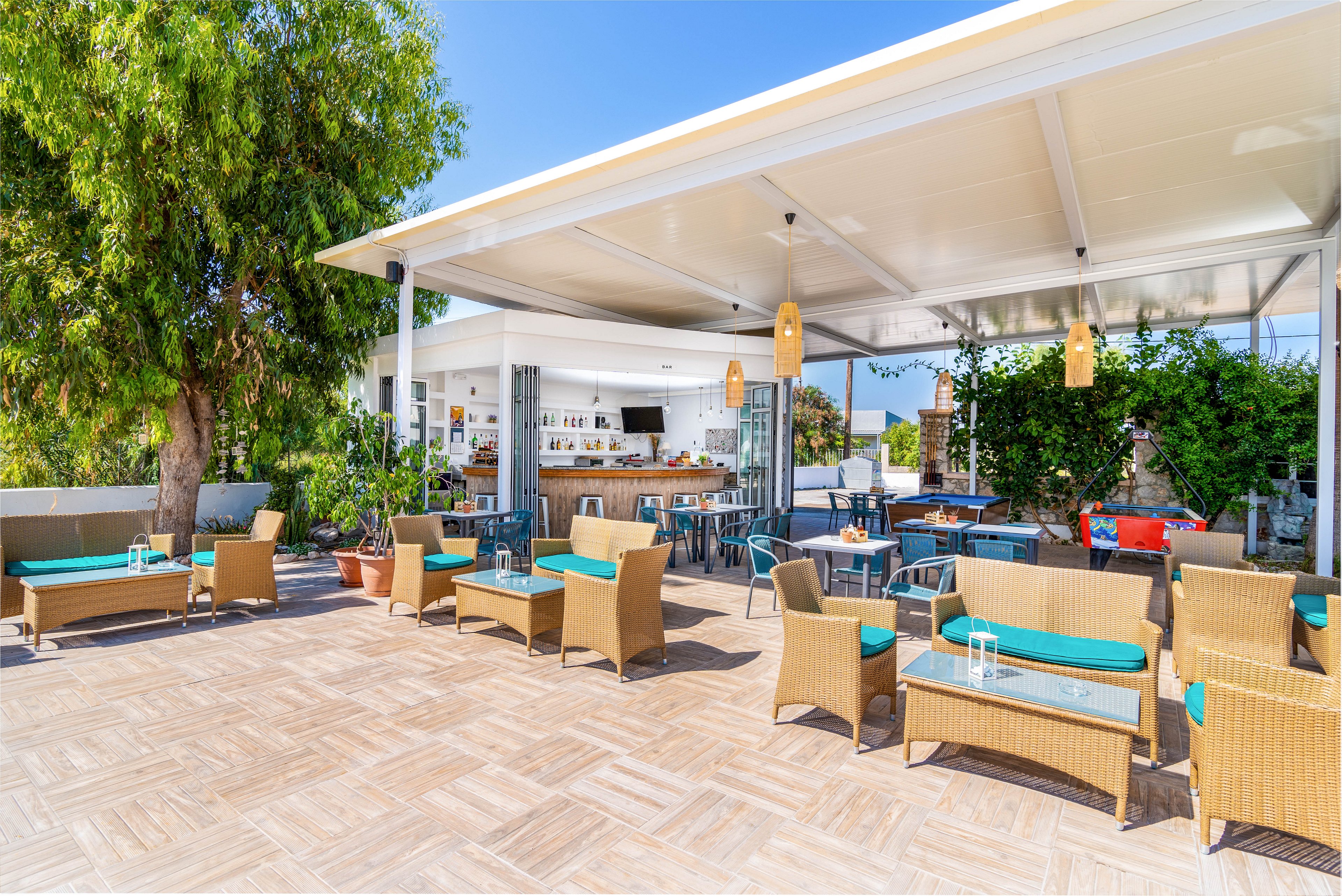 A spacious outdoor cafe features wicker chairs and sofas with turquoise cushions arranged under a modern white canopy. The setting is bright and airy, with potted plants and trees adding greenery to the relaxed atmosphere. The open bar area in the background is equipped with stools and visible shelving, while hanging pendant lights provide a stylish touch. The scene is sunlit, evoking a casual and inviting mood, ideal for social gatherings or relaxation.