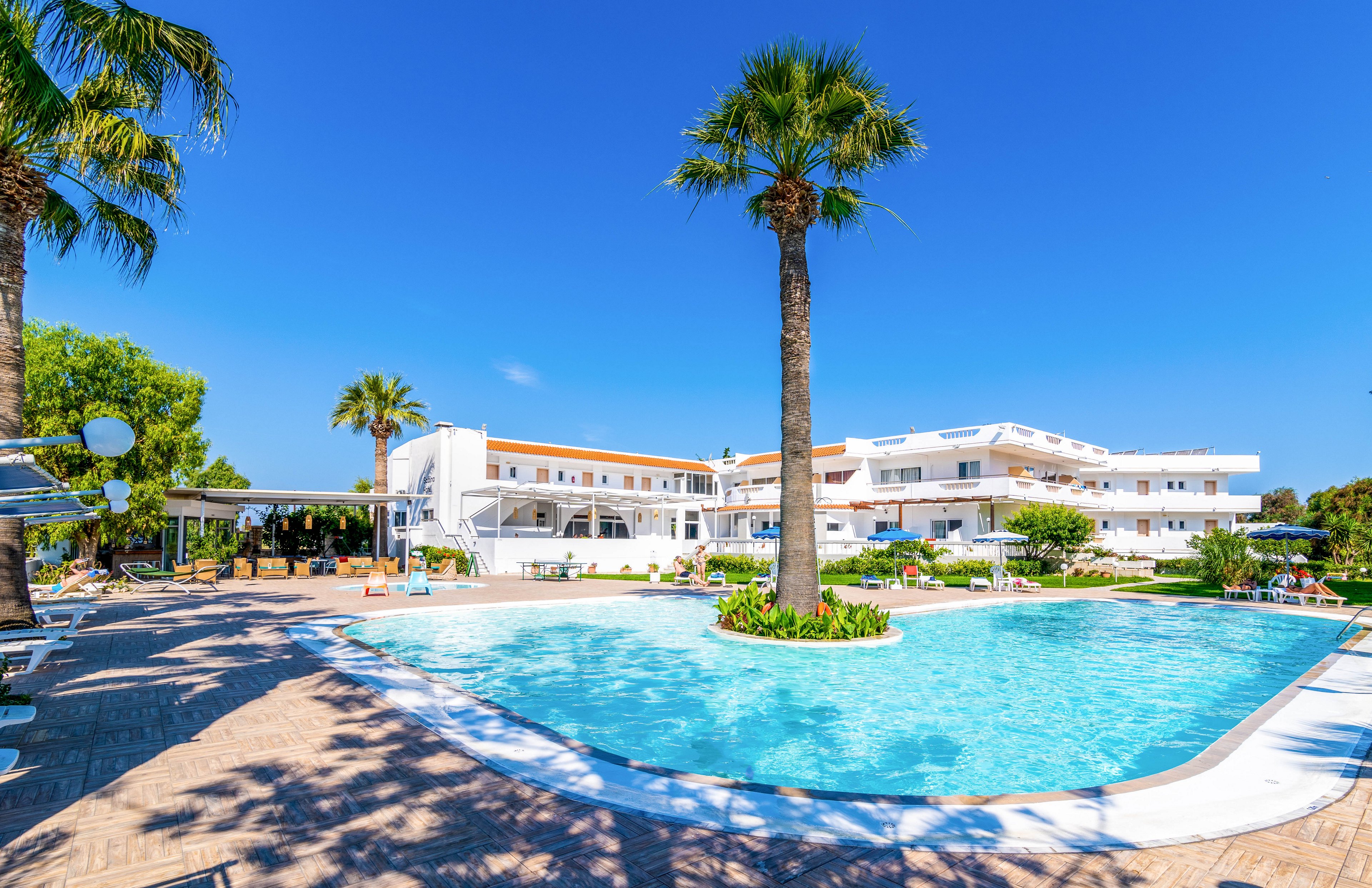 A large outdoor swimming pool is the central focus, surrounded by palm trees and sun loungers. The setting is a modern white hotel complex under a clear blue sky, evoking a relaxing vacation atmosphere. The bright sunlight and vibrant colors create a cheerful and inviting mood, with no visible people or text in the scene.