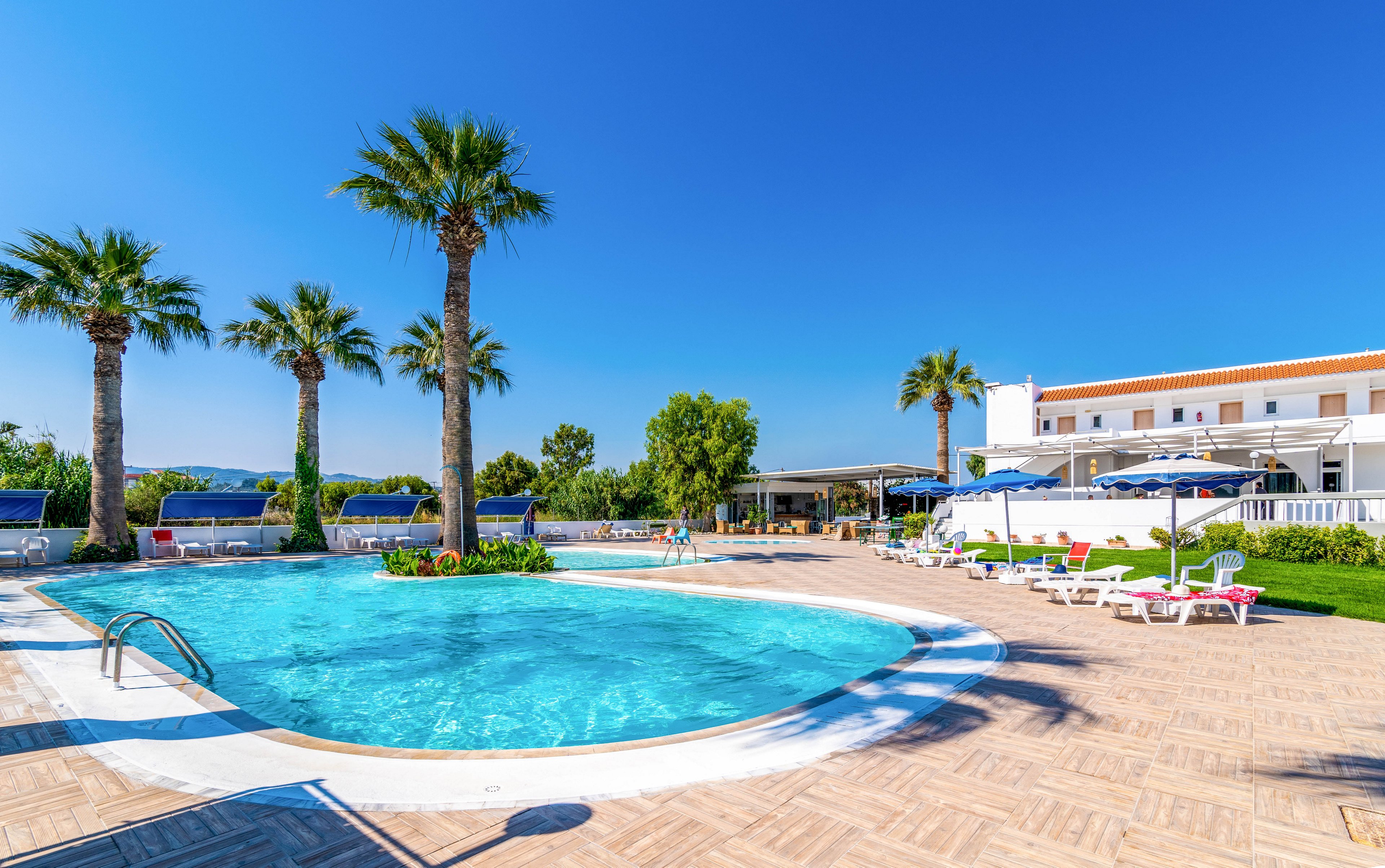A bright outdoor swimming pool is the main focus, surrounded by palm trees and sun loungers under a clear blue sky. The setting appears to be a resort or hotel with white buildings and shaded seating areas. The scene is tranquil and inviting, with a vibrant blue palette and a relaxed vacation atmosphere. No visible text or numbers are present in the image.