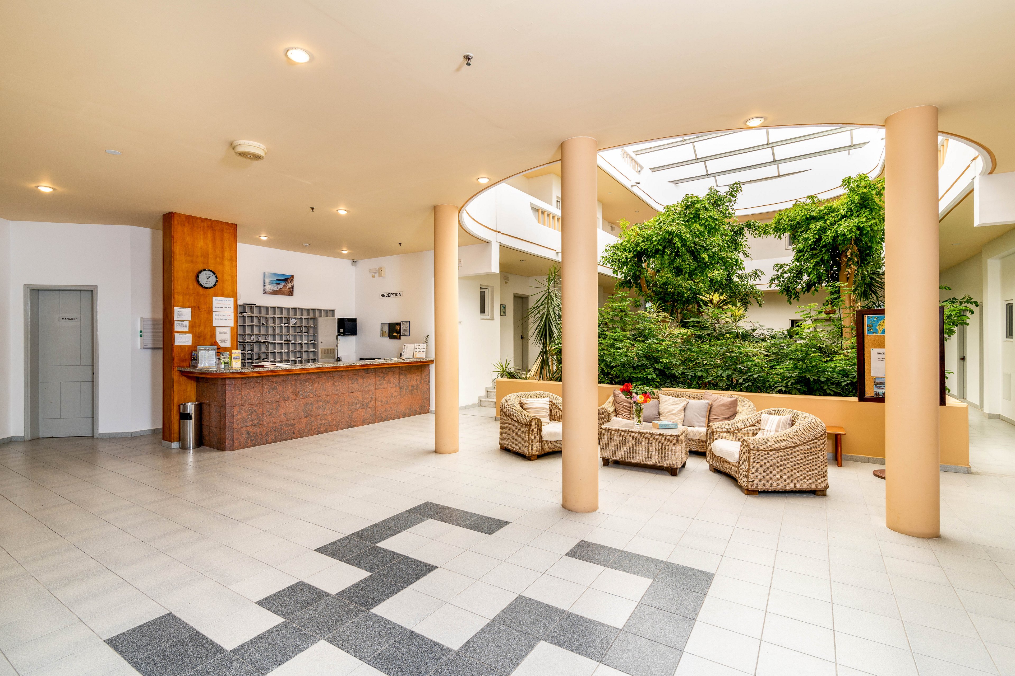 A spacious hotel lobby features a reception desk, comfortable wicker seating, and a lush indoor garden under a skylight. The setting is bright and welcoming, with neutral tones and natural light enhancing the open atmosphere. A clock and key organizer are visible behind the reception, and the tiled floor adds a clean, modern touch. No people are present in the scene.