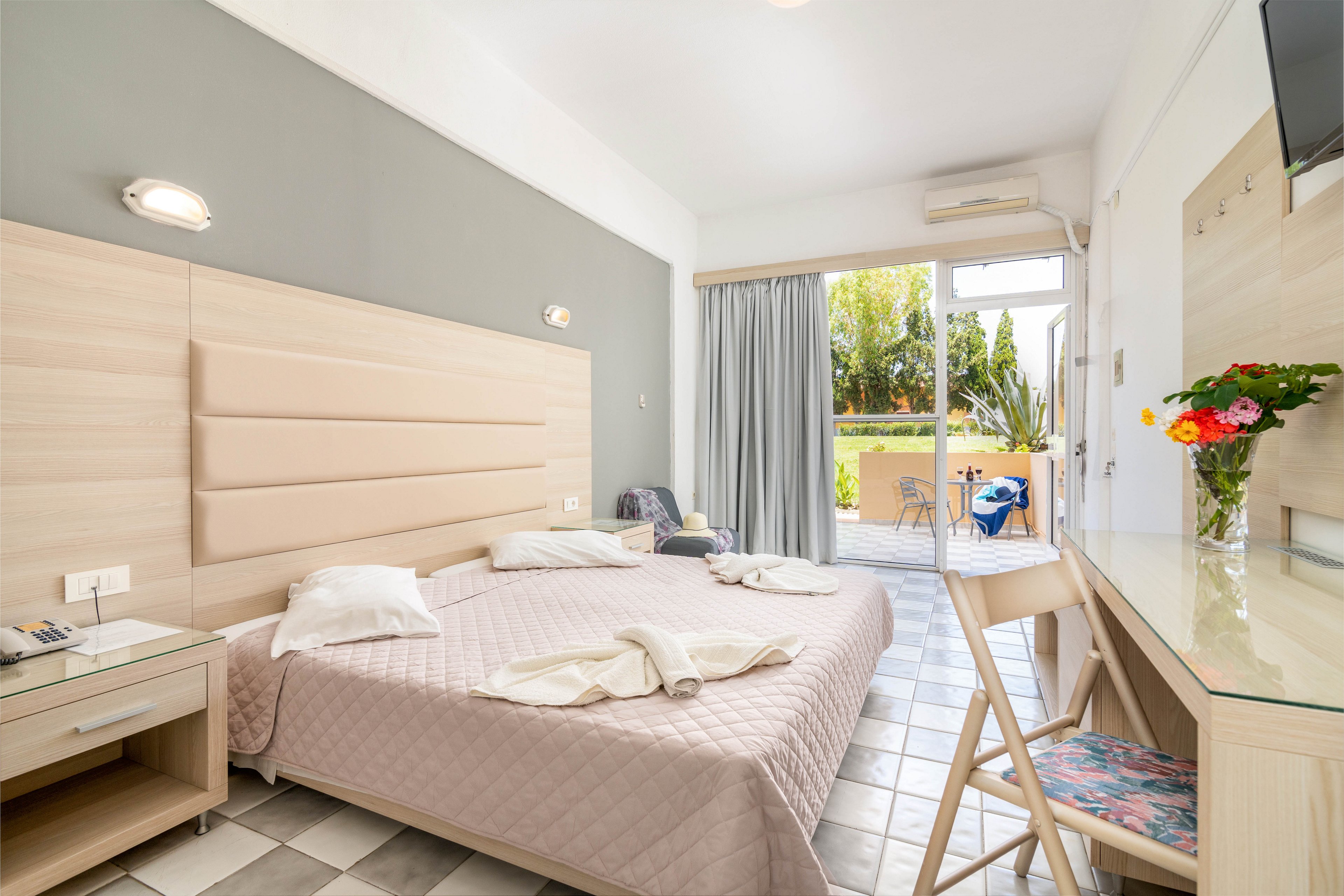 A spacious, contemporary hotel room features a double bed with folded towels and pillows, a glass-topped desk with a bouquet of flowers, and a wooden chair. The room opens to a sunlit balcony with outdoor seating, overlooking greenery. Soft neutral tones and natural light create a welcoming, relaxed atmosphere.