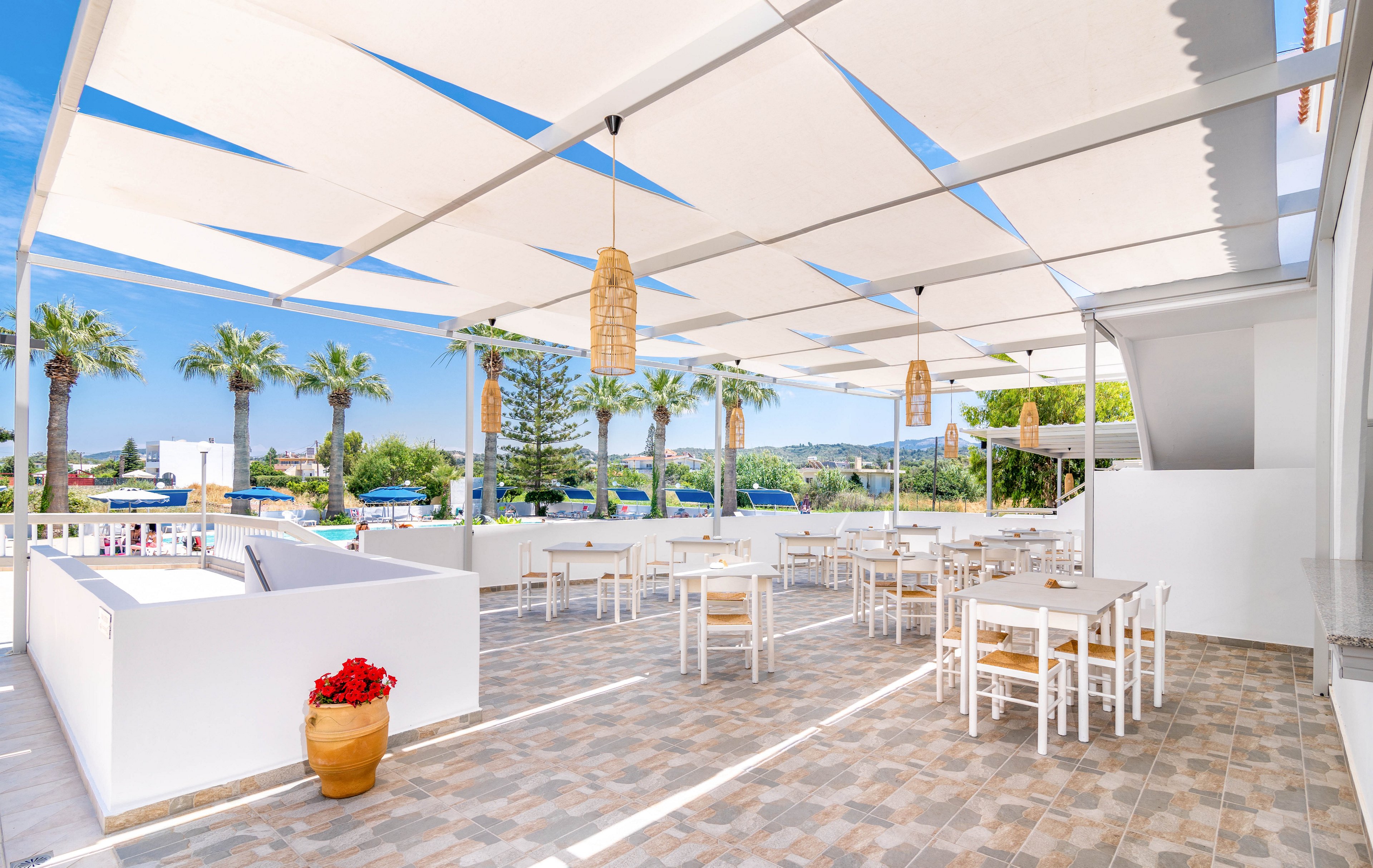 A spacious outdoor dining terrace features white tables and chairs under large geometric sunshades. The setting is bright and airy, with palm trees and a clear blue sky in the background. Natural light enhances the clean, minimalist design, while woven pendant lights add a touch of warmth. A potted plant with red flowers decorates the tiled floor, contributing to the inviting Mediterranean atmosphere.
