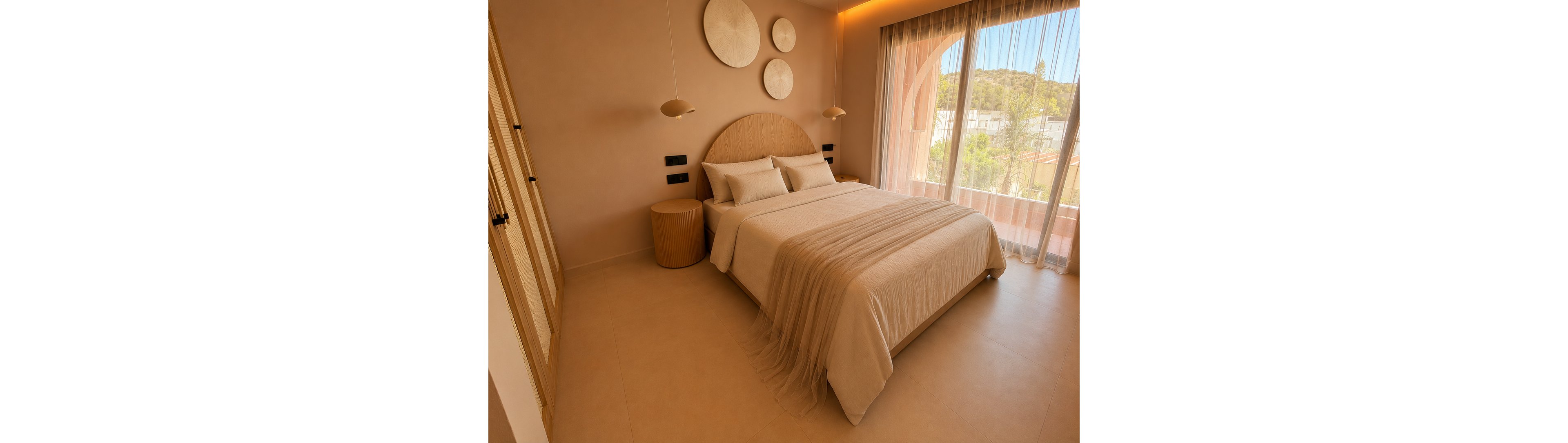 A cozy modern bedroom features a large bed with neutral beige linens and a soft throw cascading to the floor. The space is decorated in a minimalist style with a rounded wooden headboard and matching bedside table. Circular wall art and pendant lights add subtle design accents above the bed. Floor-to-ceiling sheer curtains reveal an outdoor view with greenery and architectural arches, filling the room with warm natural light.