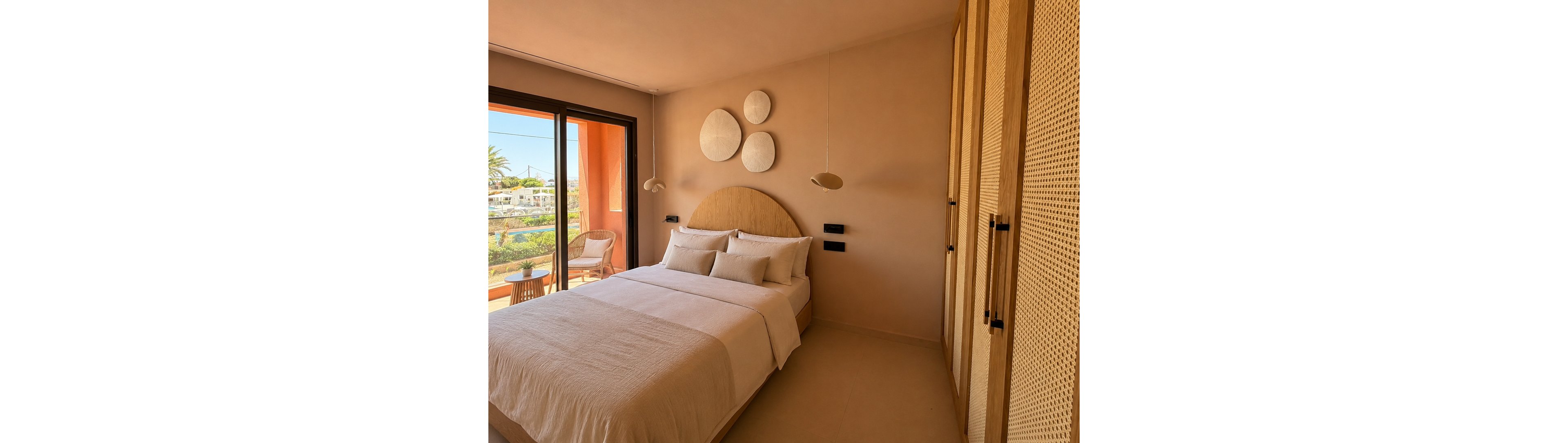 A cozy minimalist bedroom features a neatly made double bed with soft neutral bedding and multiple pillows. Warm beige walls and a wooden headboard with circular wall art create a calm, contemporary atmosphere. Sliding glass doors open to a balcony with a chair and outdoor view, allowing natural light to fill the room. Built-in wardrobes with textured doors and modern pendant lights complete the serene interior design.