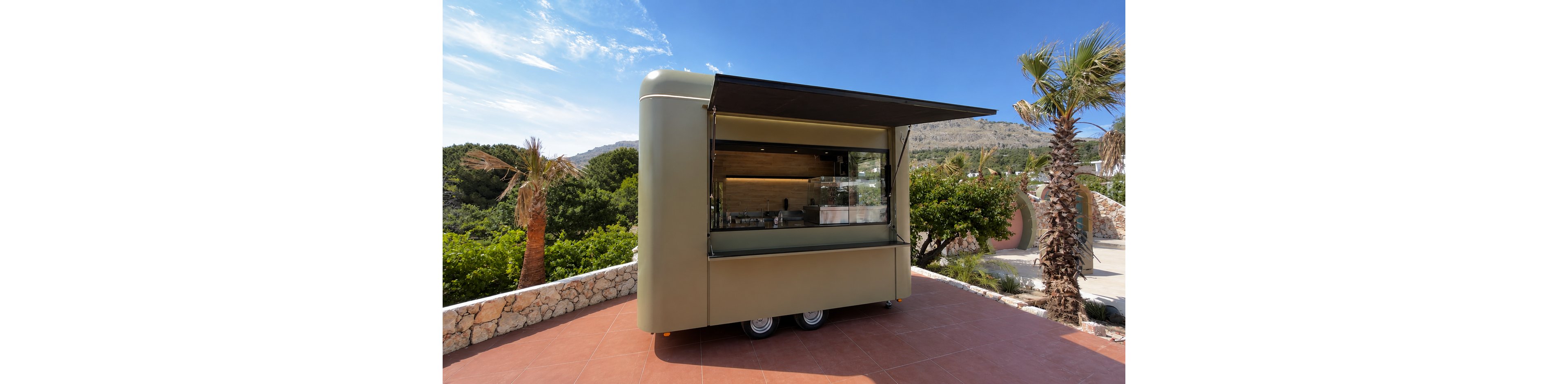 A sleek, modern food truck stands parked on a tiled terrace in a sunny outdoor setting. The service window is open, revealing a minimalist wooden interior and glass display cases. Lush green trees, stone walls, and distant hills create a relaxed resort atmosphere. Bright daylight and clear blue skies emphasize the warm, inviting vacation mood.