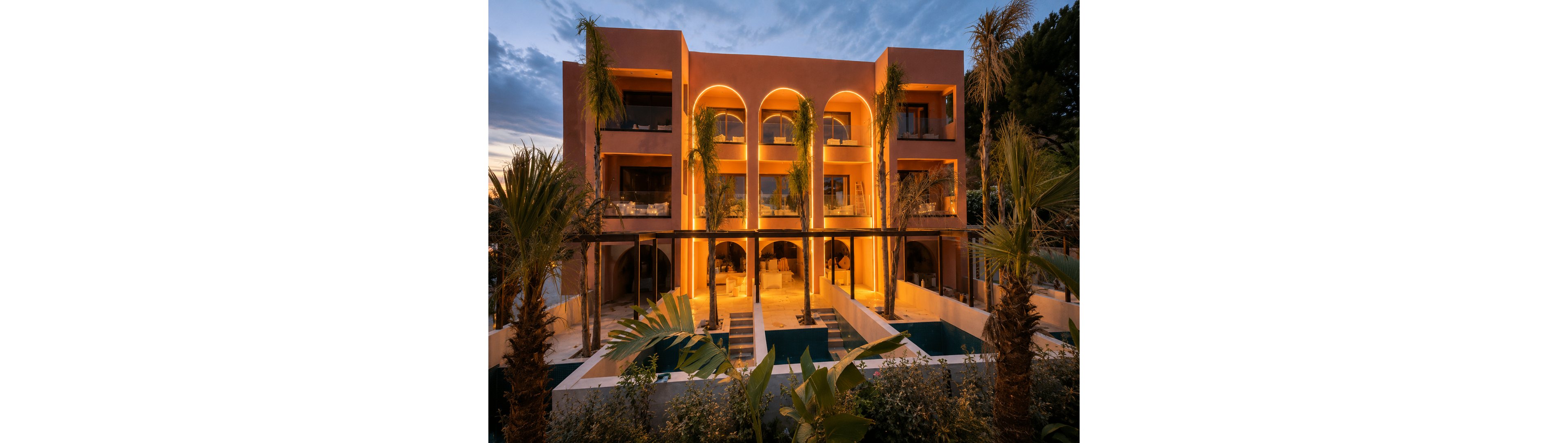 A contemporary multi-story hotel glows warmly at dusk, with large arched openings and balconies facing a landscaped courtyard. The building’s exterior lighting creates a golden contrast against the deepening blue evening sky. Palm trees and lush greenery frame a series of narrow pools or water features in the foreground. The scene evokes a sense of luxury, relaxation, and resort-style living.