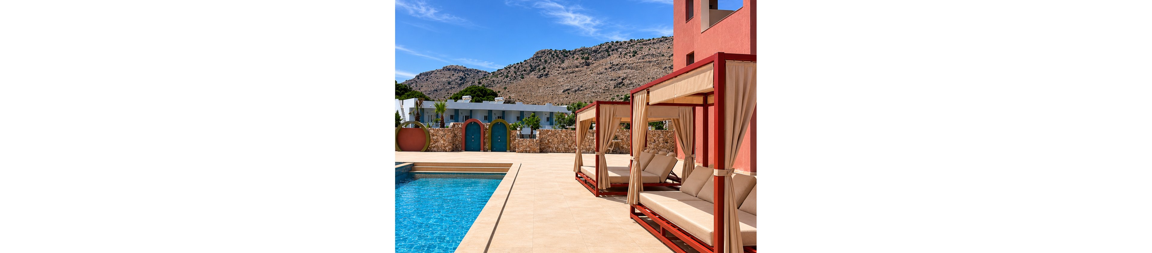 Outdoor cabanas with cushioned loungers line the edge of a clear blue swimming pool at a modern resort. The scene is set against a backdrop of rocky hills and a bright blue sky. Contemporary buildings and stone walls frame the pool area, creating a clean, geometric composition. Warm beige tiles and red cabana frames contrast with the turquoise water for a relaxed, upscale vacation atmosphere.